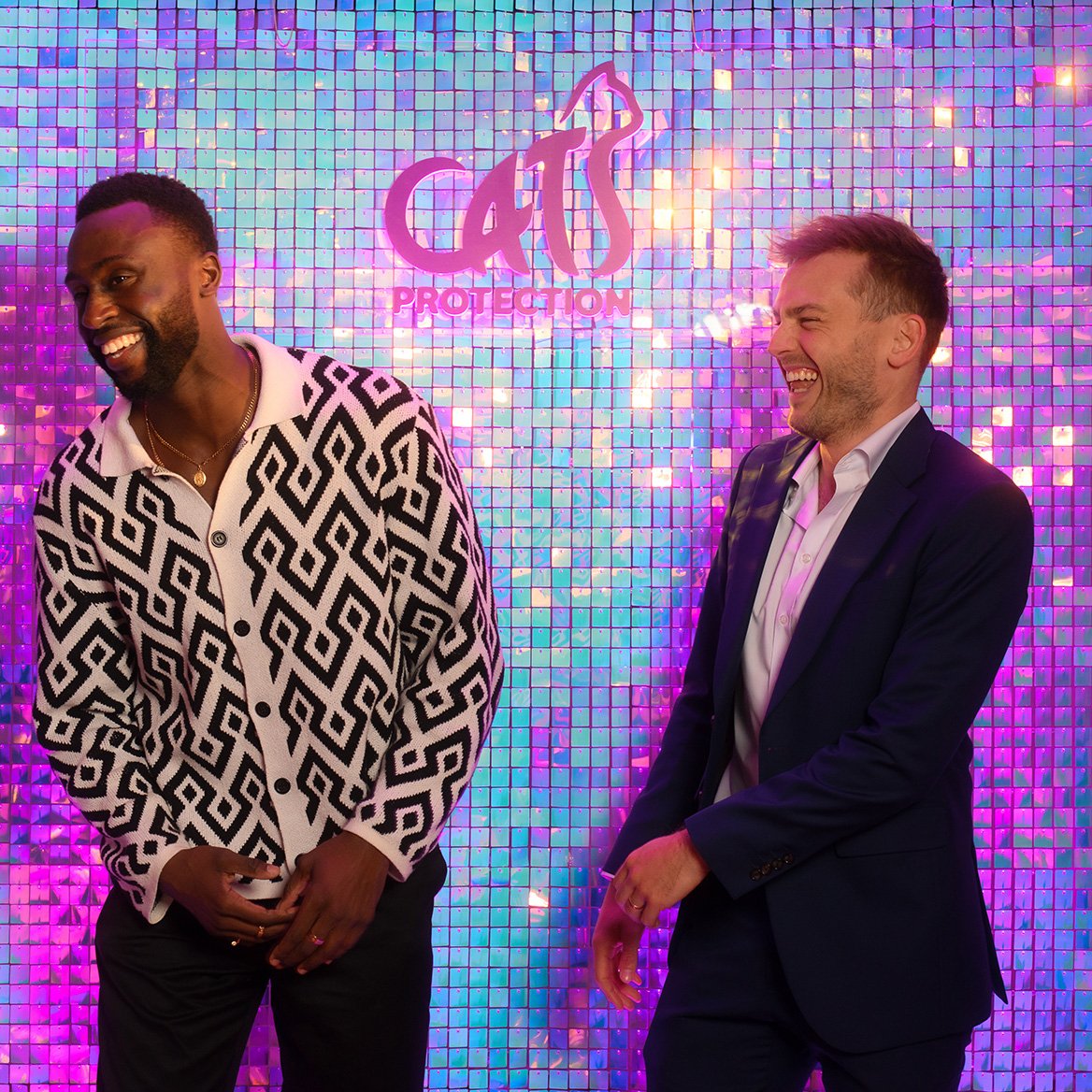 Two men stand in front of a colorful, sparkly backdrop with the words 'Arts Protection' in pink and purple. They are laughing and appear to be having a good time.