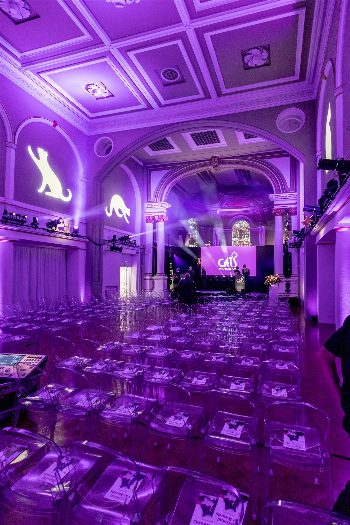 A large event hall illuminated with purple lighting, decorated with elegant architecture, including arched windows, columns, and detailed ceiling moldings. The hall has rows of transparent chairs, and a stage with screens displaying the text 'CAT PROTECTION' and silhouettes of cats, indicating a feline advocacy event.