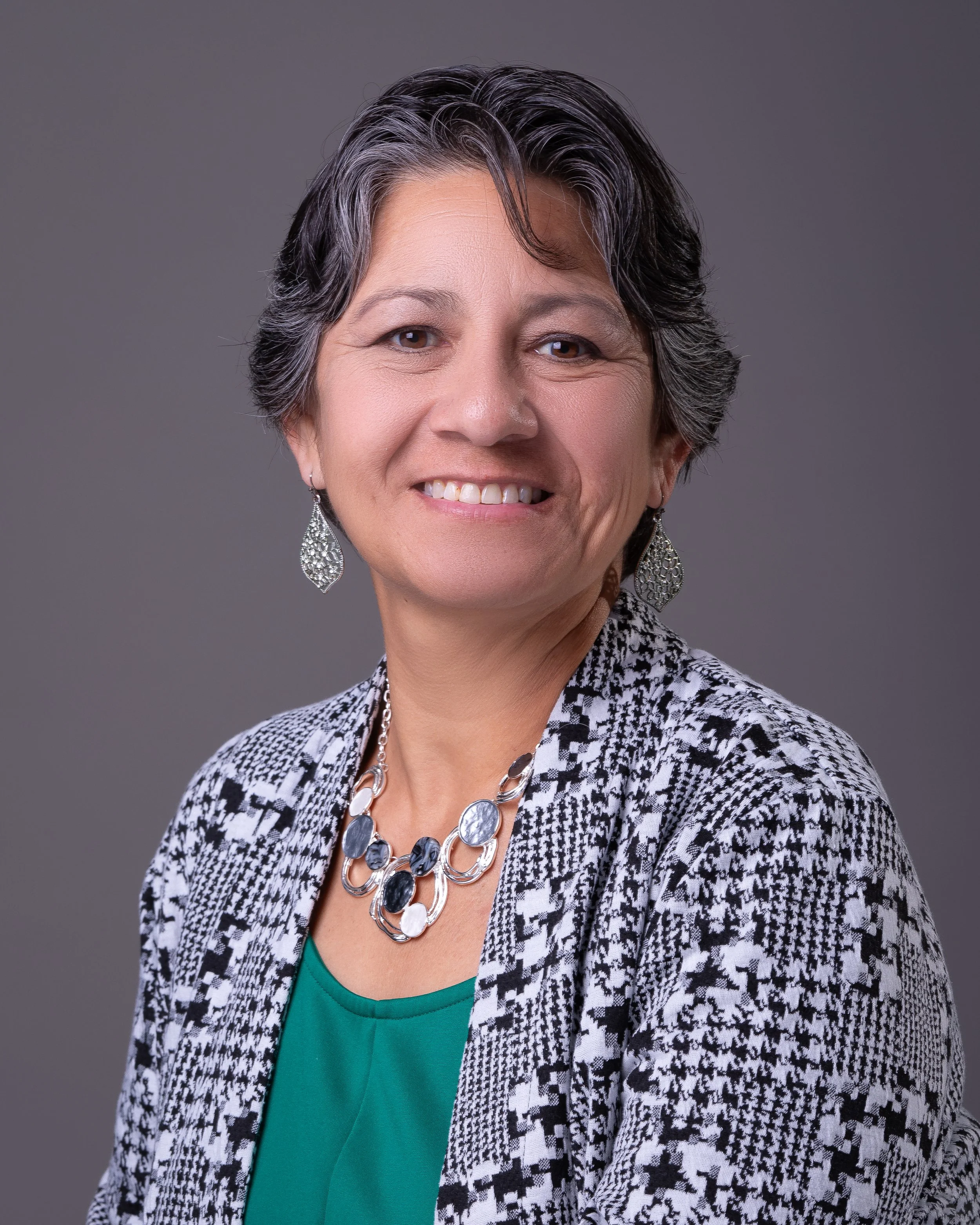 Close-up of a middle-aged woman with short, dark, and gray hair, smiling, wearing a patterned blazer with a necklace and earrings against a neutral background.