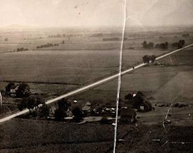 Black and white aerial photo of a rural landscape with fields, a farm, and a train traveling across the landscape.