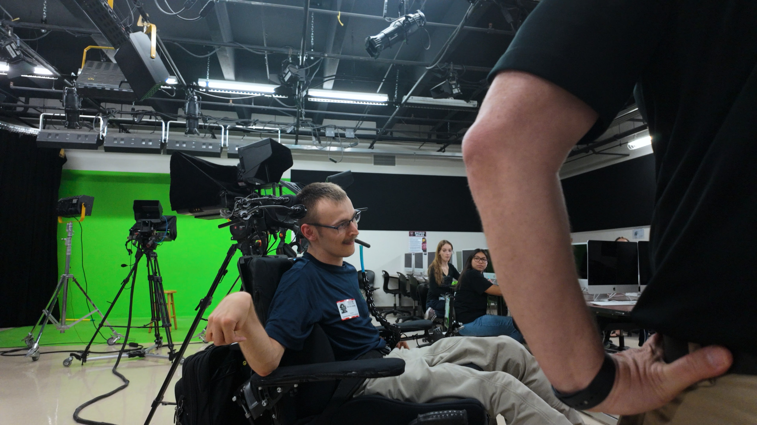 A man sitting in a studio with camera equipment and a green screen behind him, while two women observe and an individual stands partially in view.