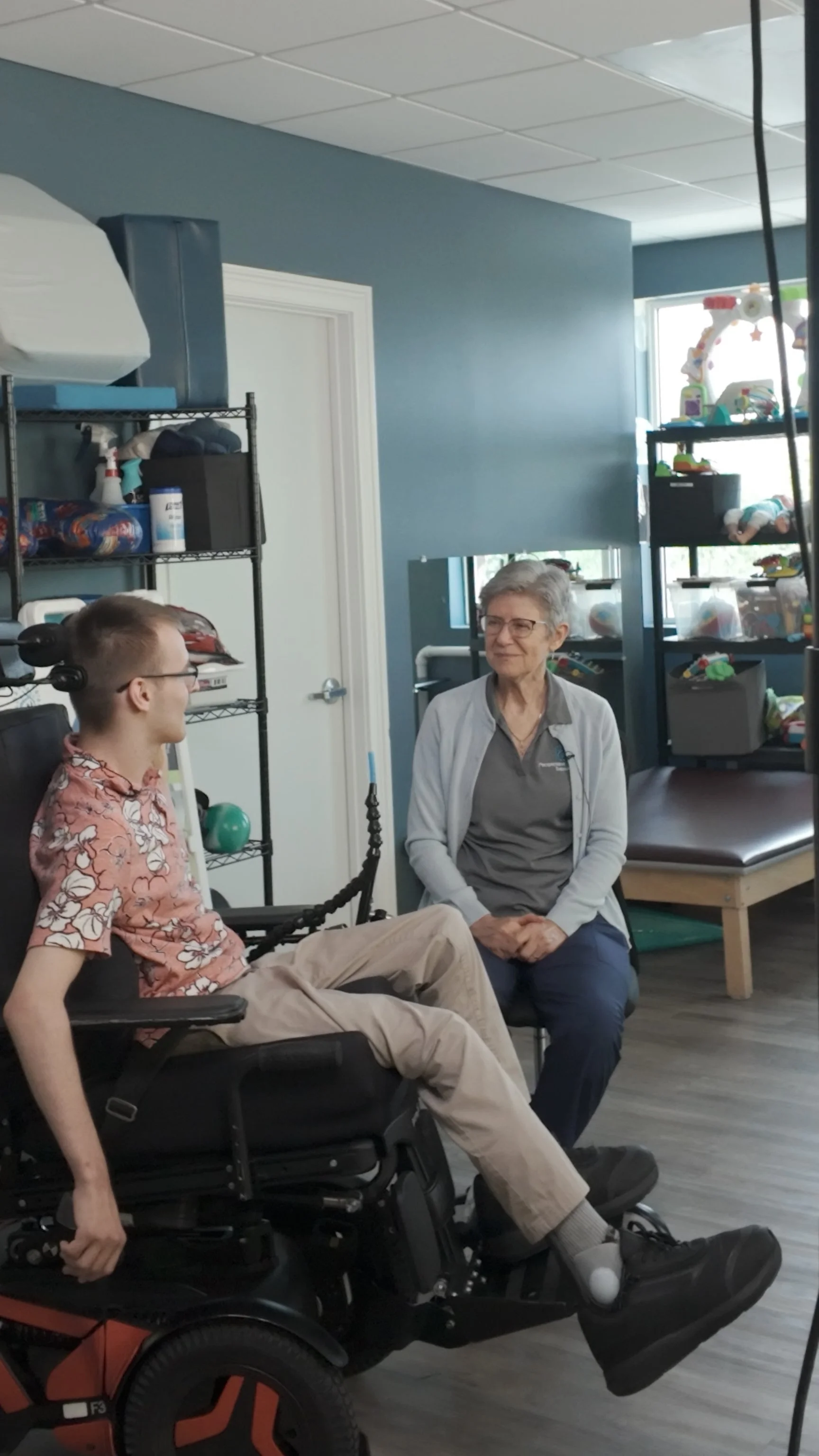 A young man in a wheelchair and an older woman sitting on a chair in a room, engaging in conversation, with shelves of toys and a therapy table in the background.