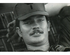 A black-and-white photo of a young man with a mustache, wearing a baseball cap and lying down on a pillow.