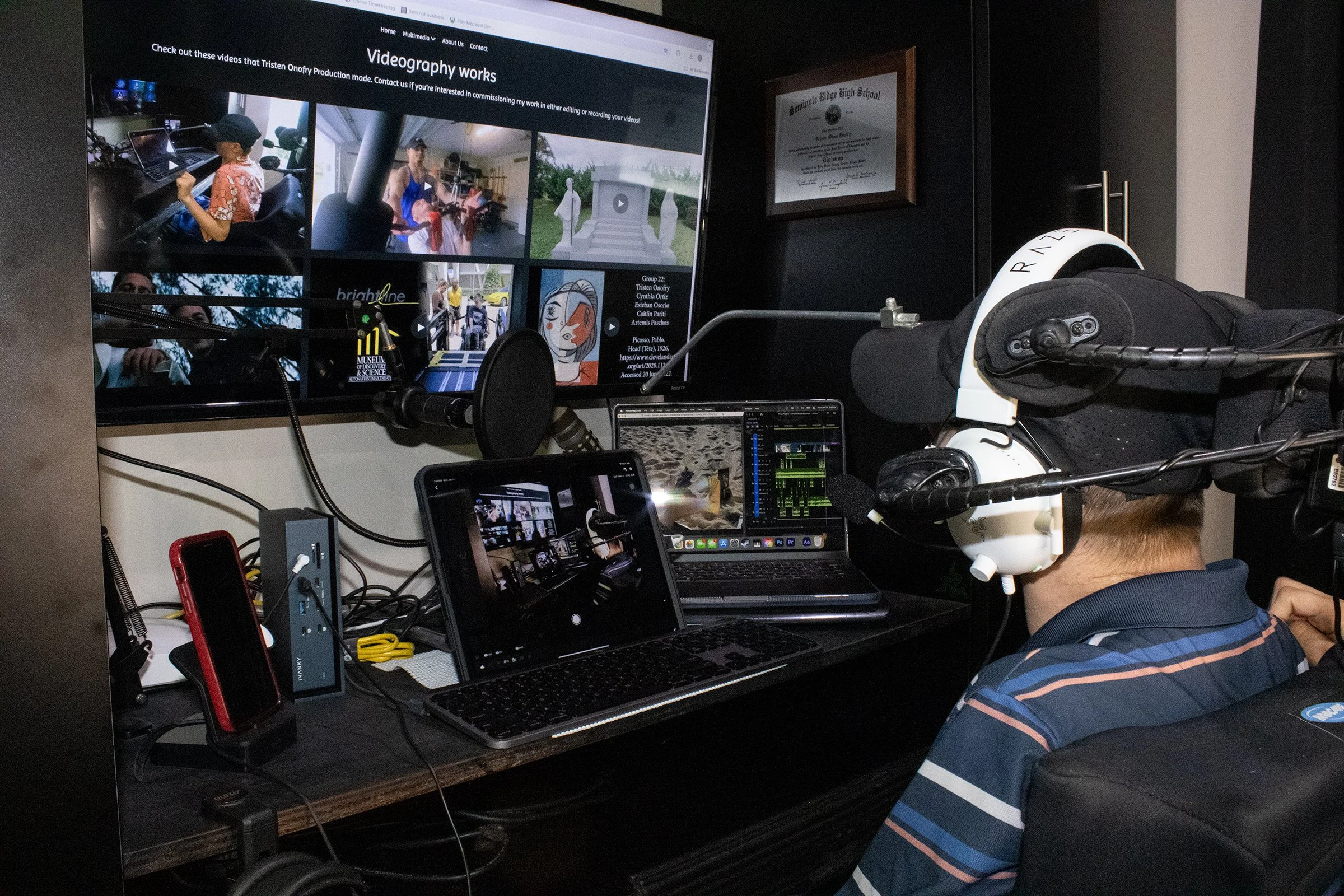 A person wearing headphones is seated at a desk with multiple computer screens, laptops, and recording equipment, working on video editing or production in a dimly lit room.
