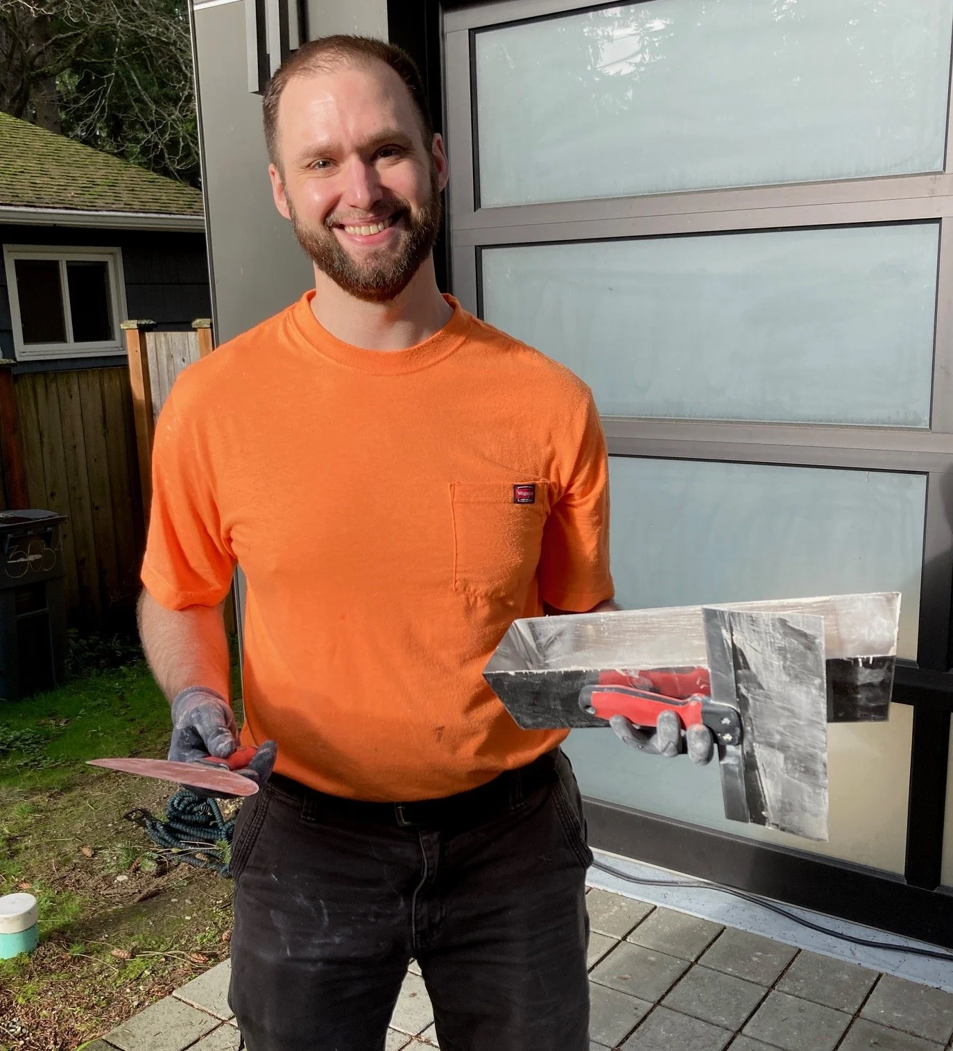 A man with a beard and short hair smiling while holding a trowel and a float, wearing an orange shirt and black pants, standing outside in front of a gray garage door.