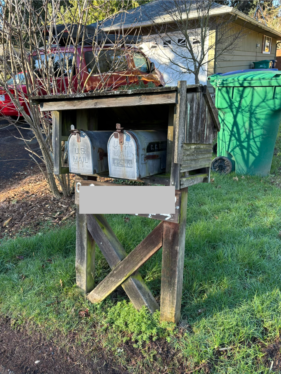 A wooden mailbox post with two metal mailboxes located in a yard, nearby a bush and under a tree. In the background, there's a house, a green trash bin, and some parked cars.
