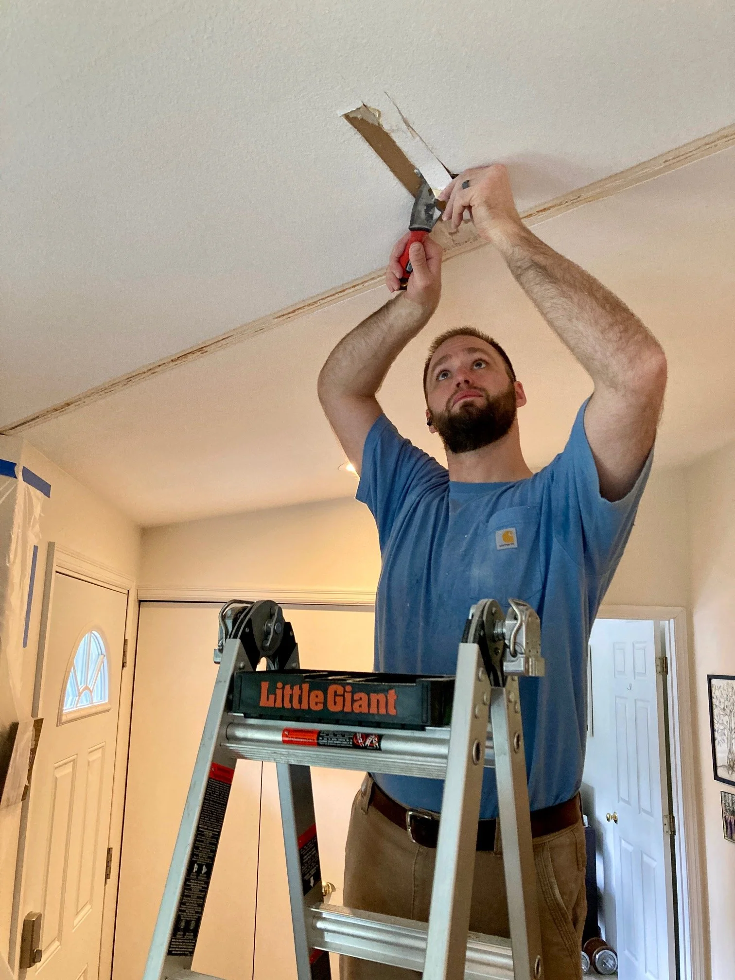 A man standing on a Little Giant step ladder, working on the ceiling with a putty knife, in a room with a front door, closet, and doorway visible.