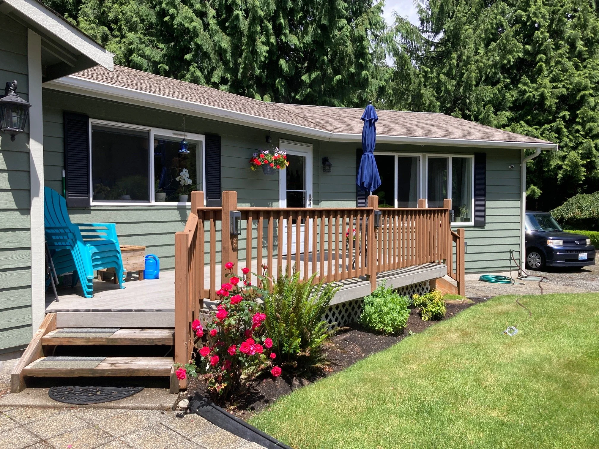 Frontyard of a house with a green exterior and a small wooden deck with a blue patio umbrella. There are pink flowers in the garden bed beside the deck, and a black car parked in the driveway. Tall trees are in the background.