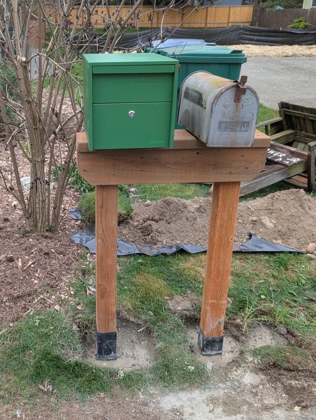 A wooden mailbox post with a green mailbox and an old metal mailbox on top, placed outdoors in a yard with dirt, grass, and bushes.