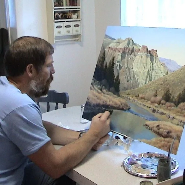 Mike Stinnett sits at a table painting a landscape featuring Cathedral Rock using acrylic paints.