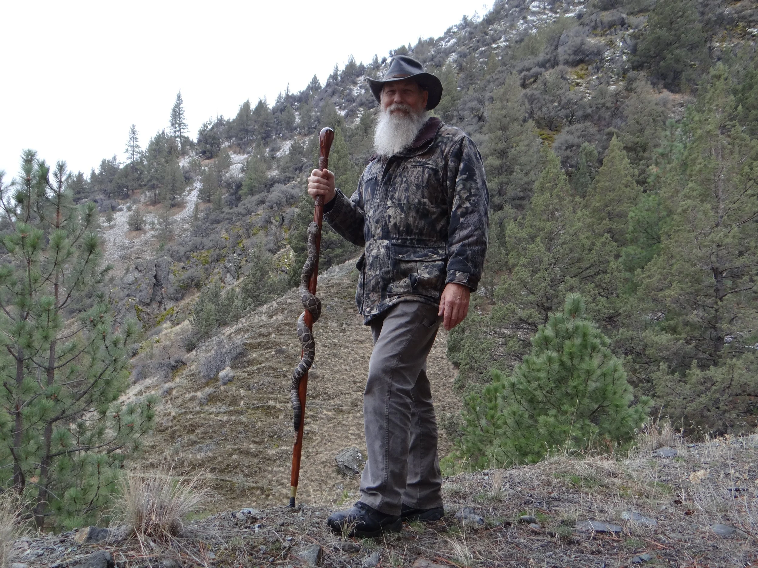 Mike Stinnett stands holding one of his wood carved rattlesnake walking sticks in a rugged mountain setting.