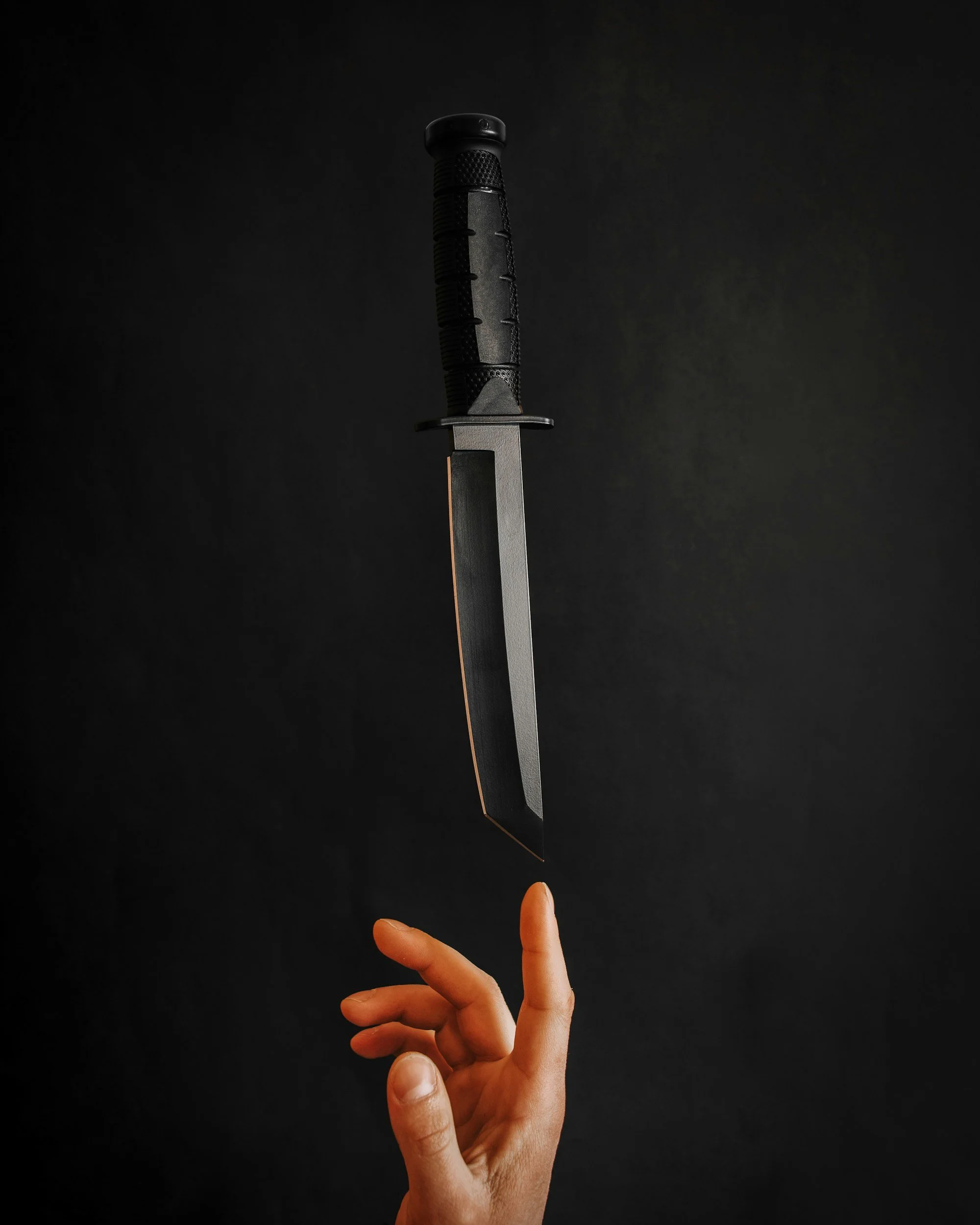 Una mano con dedos extendidos sostiene una cuchilla que parece un sastre, apuntando hacia arriba sobre fondo negro.