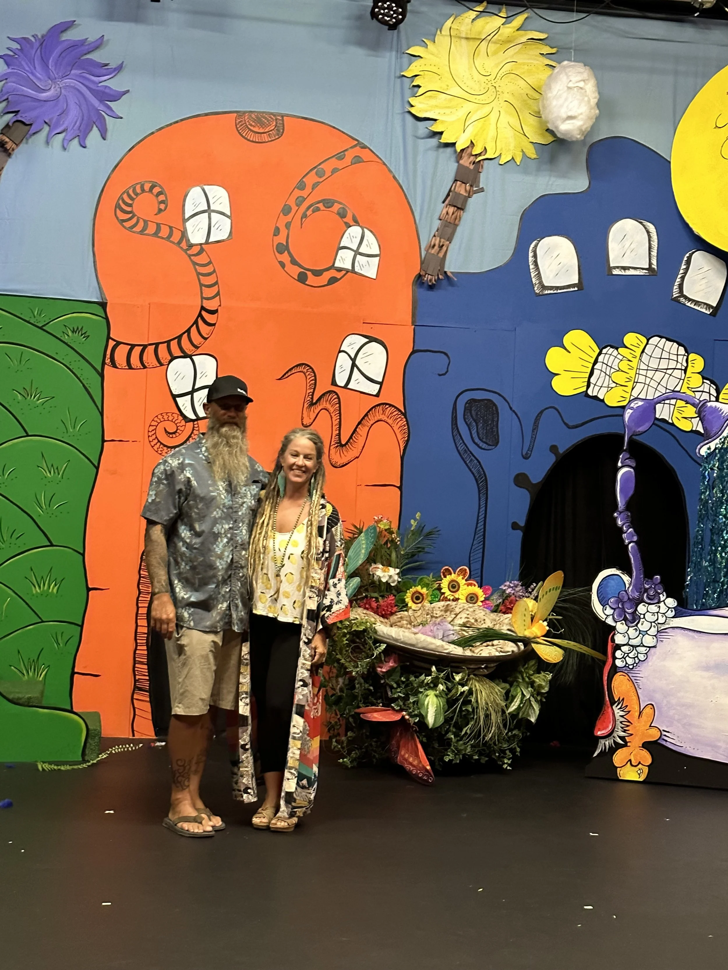 Two people standing in front of a colorful, cartoon-style backdrop featuring a large, orange snake, green hills, and abstract yellow, purple, and blue shapes with flowers and leaves at the base.