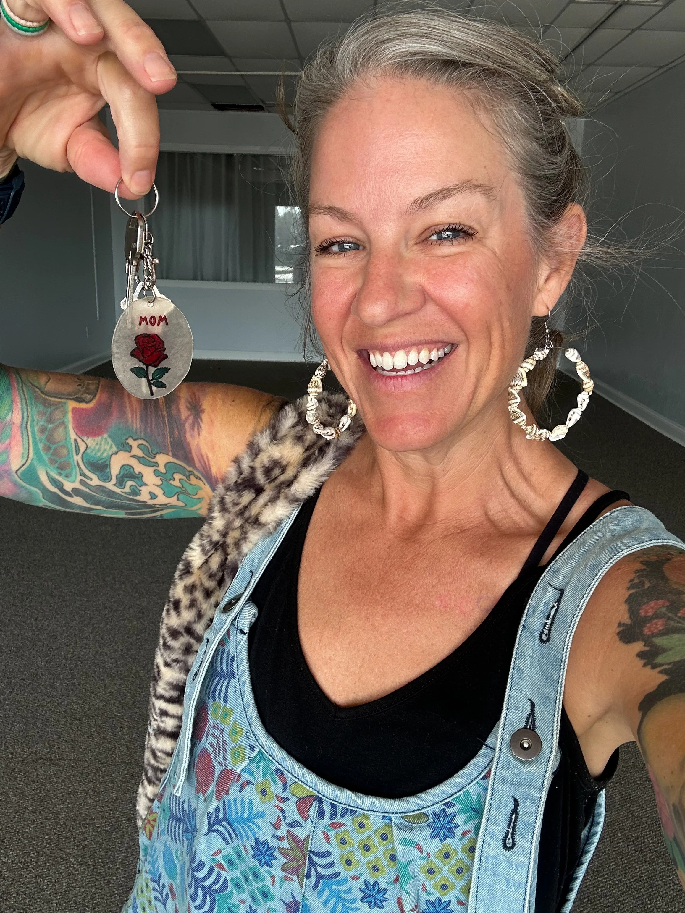 A smiling woman with tattoos on her arm and earrings, holding a keychain with a red rose and the word 'MOM' on it, in an indoor setting.