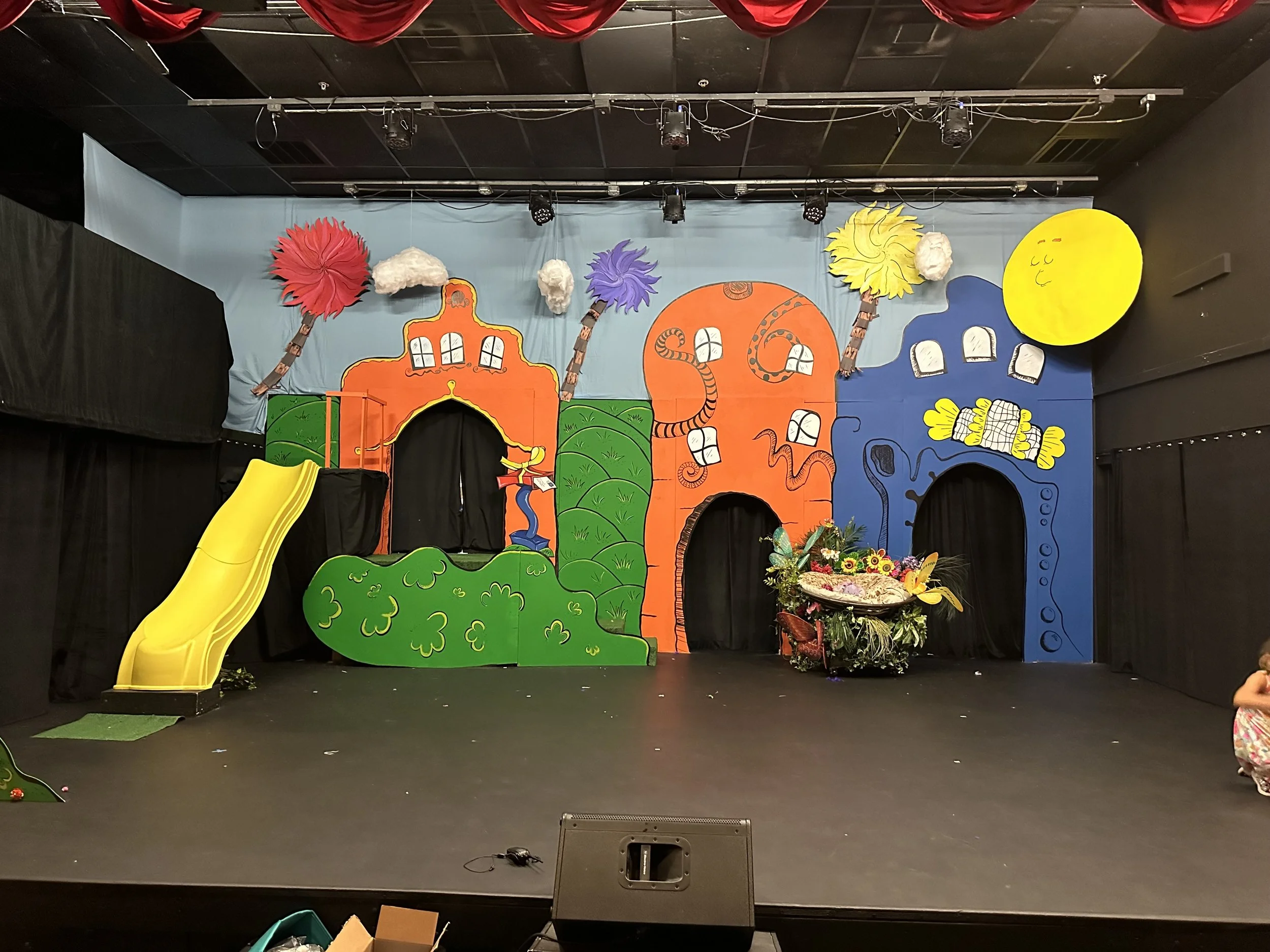 Colorful stage set with cartoon-style trees, buildings, flowers, and a yellow slide, decorated for a children's performance or event.