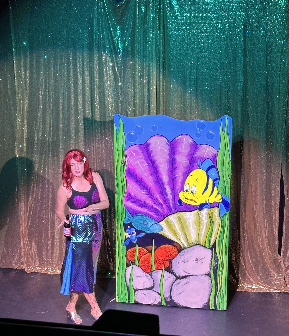 A woman with red hair and fishnet stockings stands on a stage next to a colorful painting of a cartoon underwater scene featuring a yellow fish with blue stripes, purple coral, and green seaweed, with a shimmering gold curtain in the background.