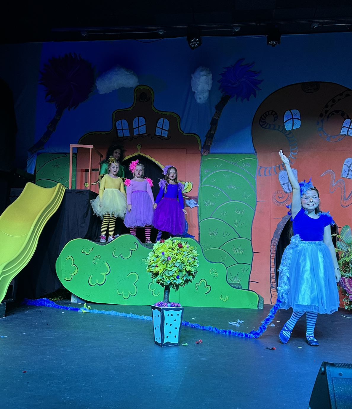 Children performing a play on stage with colorful backdrop, some kids are in bright costumes and one girl in a blue dress with a blue headband is raising her hand.