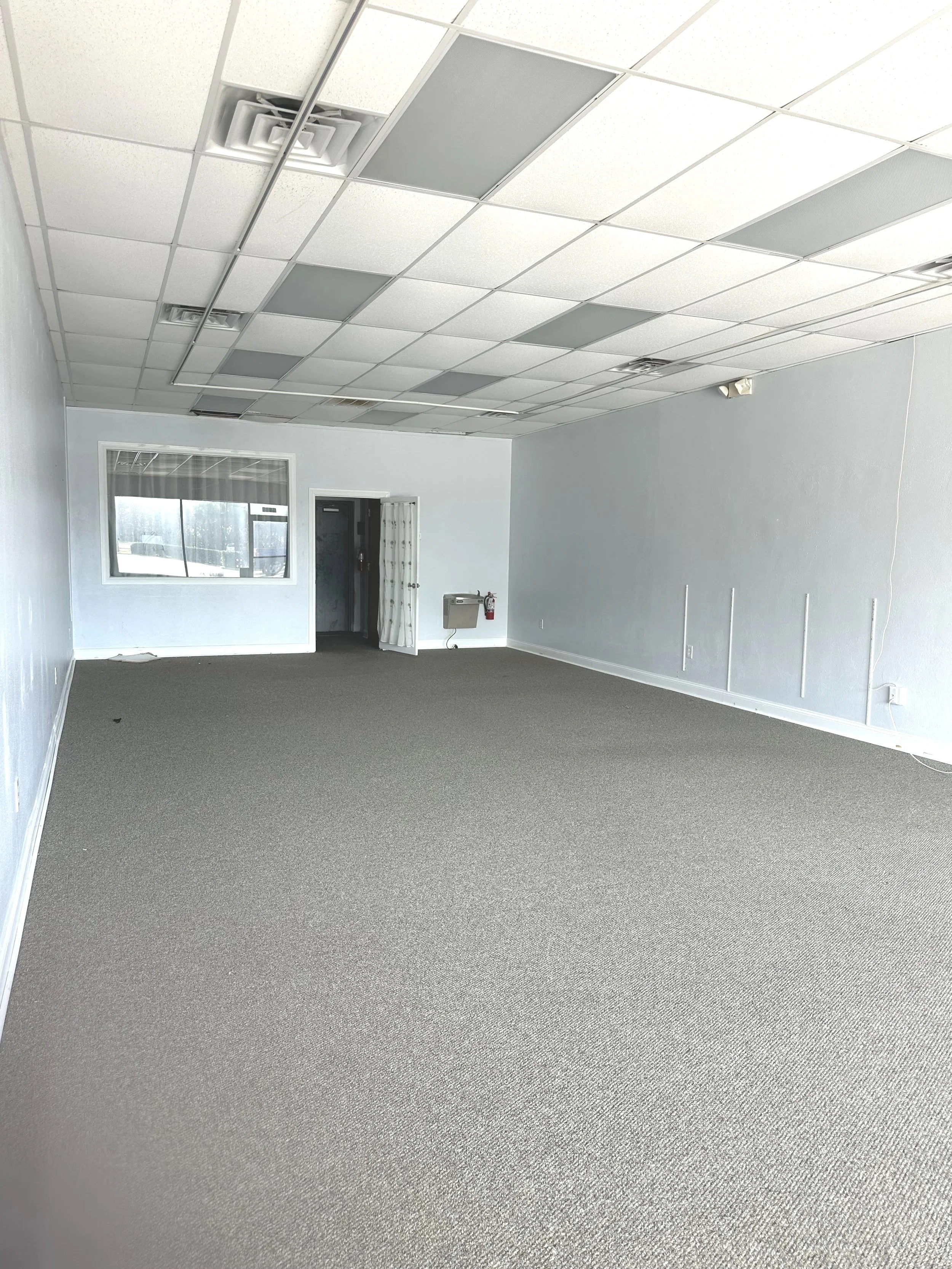 Empty room with gray carpet, white walls, and a ceiling with square panels and fluorescent lights overhead.
