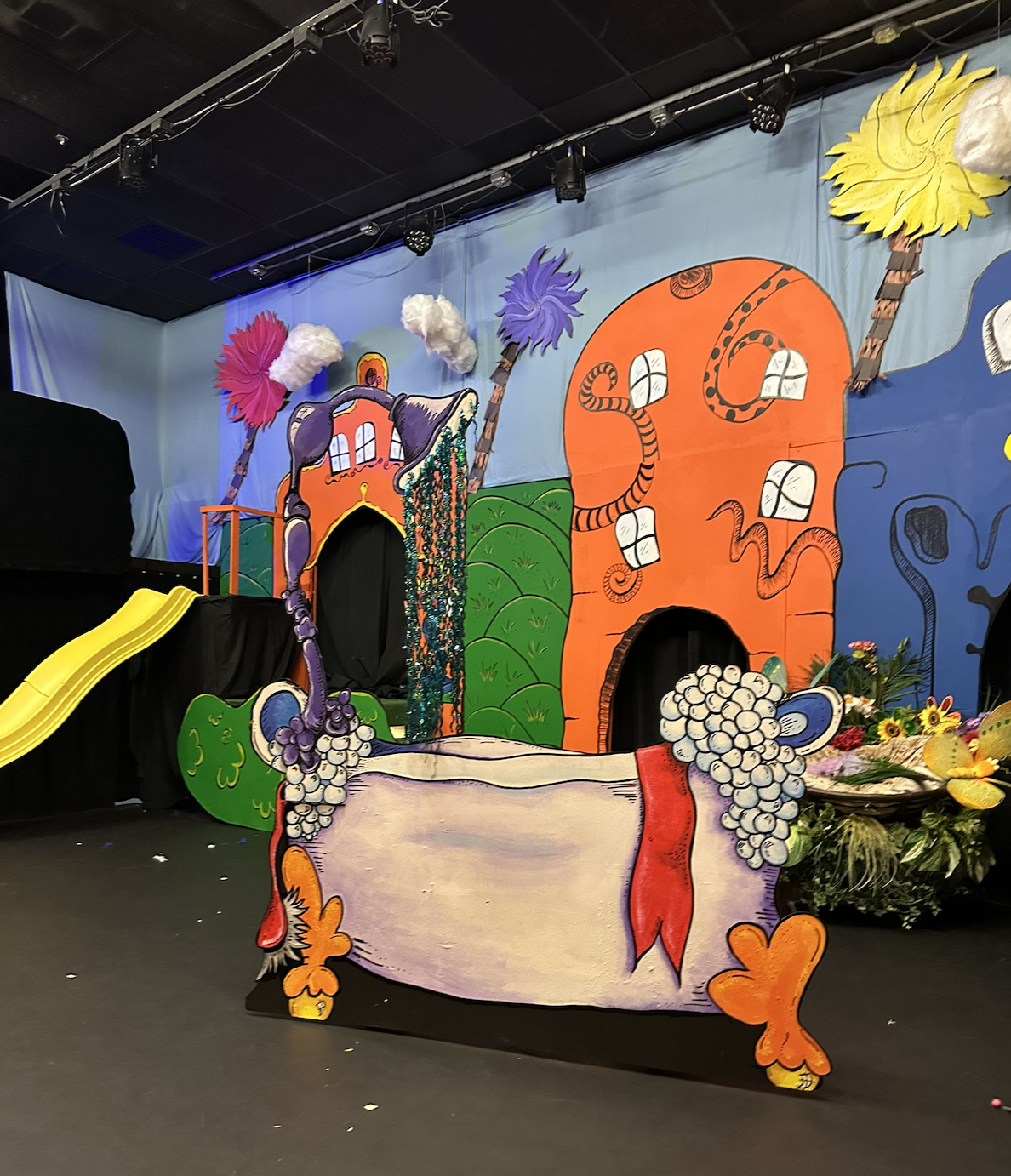 Colorful stage set with cartoon-style buildings, trees, clouds, and a large decorative wine jug with grapes in the foreground, indicating a festive or theatrical event.