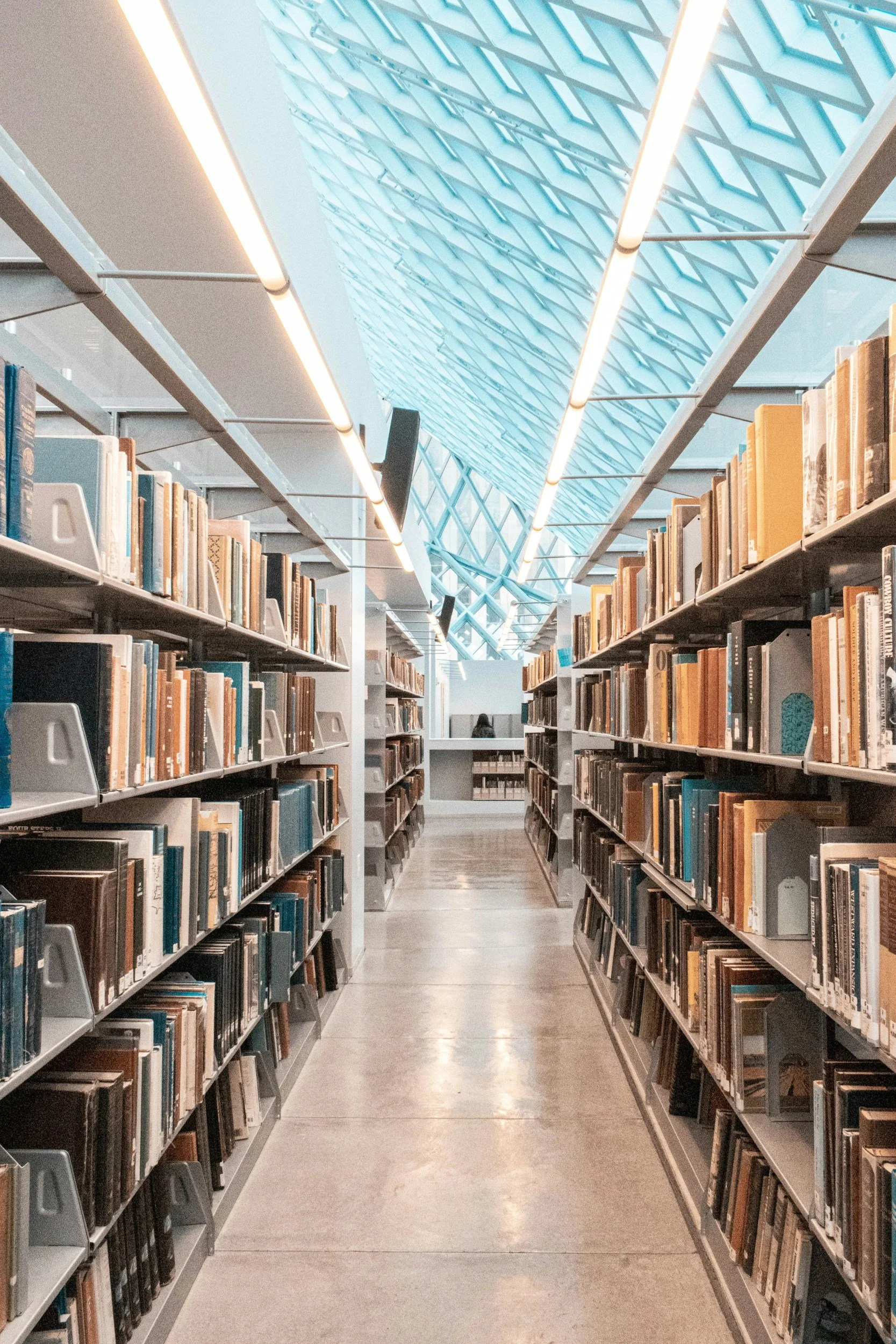 Aisle in a modern library with bookshelves filled with books on both sides and a glass ceiling above, reflecting the architectural structure.