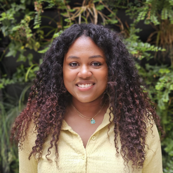 Woman with curly dark hair and a gold shirt smiling outdoors with green plants in the background.