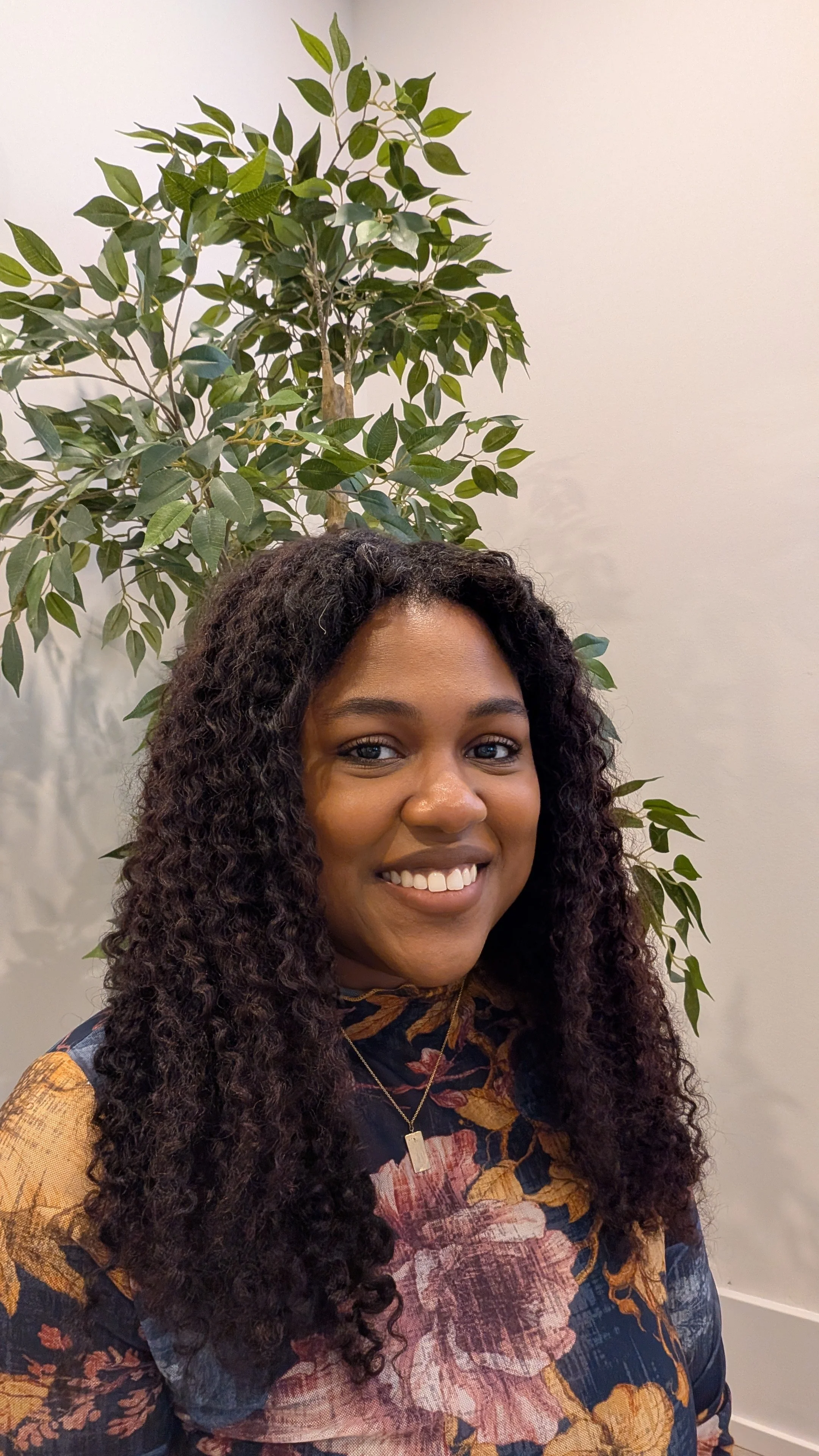 A woman with long, curly dark hair smiling at the camera, standing in front of a leafy green plant and a plain white wall.