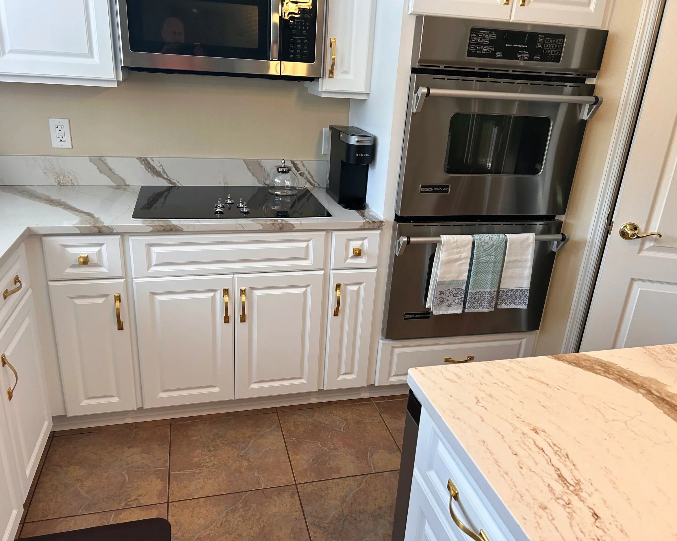 White kitchen cabinets with gold handles, a microwave, a stove with black cooktop, a Keurig coffee maker, a clear glass jar, towels hanging on a rack, and beige marble countertops with tile flooring.