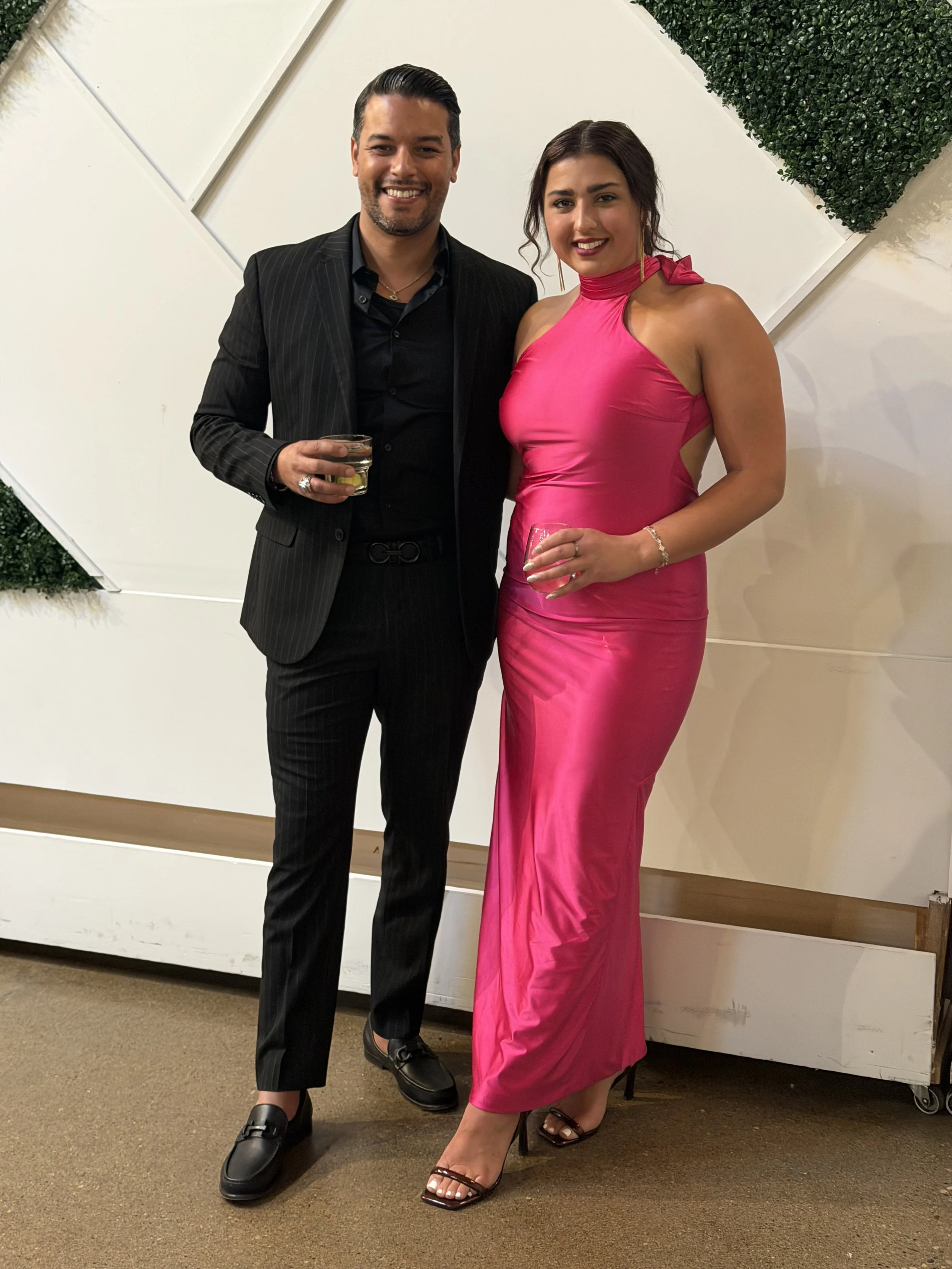 A man and a woman standing together, smiling, dressed in formal attire, holding drinks at an indoor event.