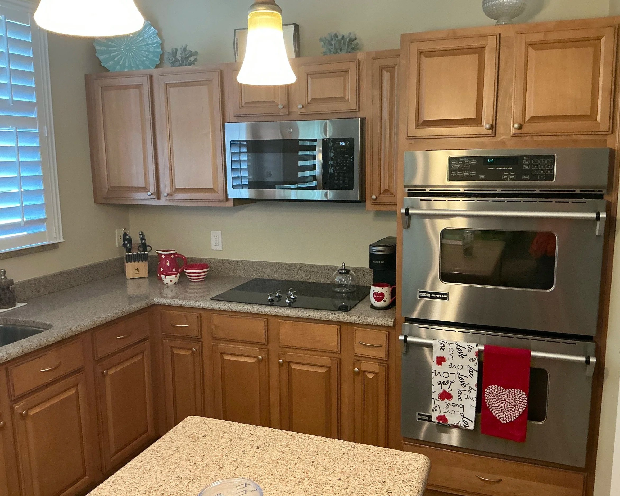 A kitchen with wooden cabinets, stainless steel microwave and oven, beige countertop, a window with white shutters, decorative dishes on top of the cabinets, and kitchen towels hanging on the oven handle with love-themed designs.