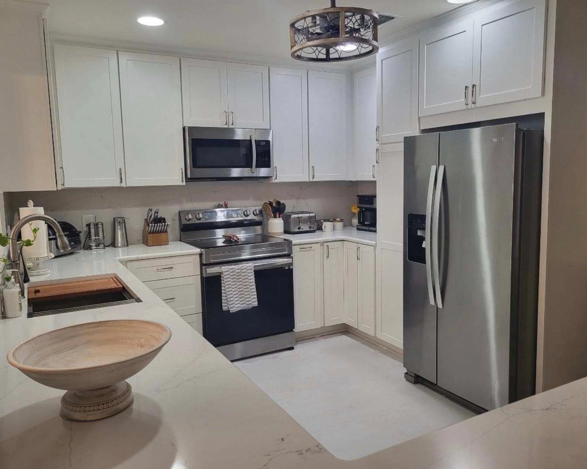 A modern kitchen with white cabinets, stainless steel appliances, and a white counter. There is a built-in microwave above the stove and a large stainless steel refrigerator. The kitchen island has a wooden bowl and a sink with a faucet. Overhead lighting and a decorative circular ceiling light fixture are visible.
