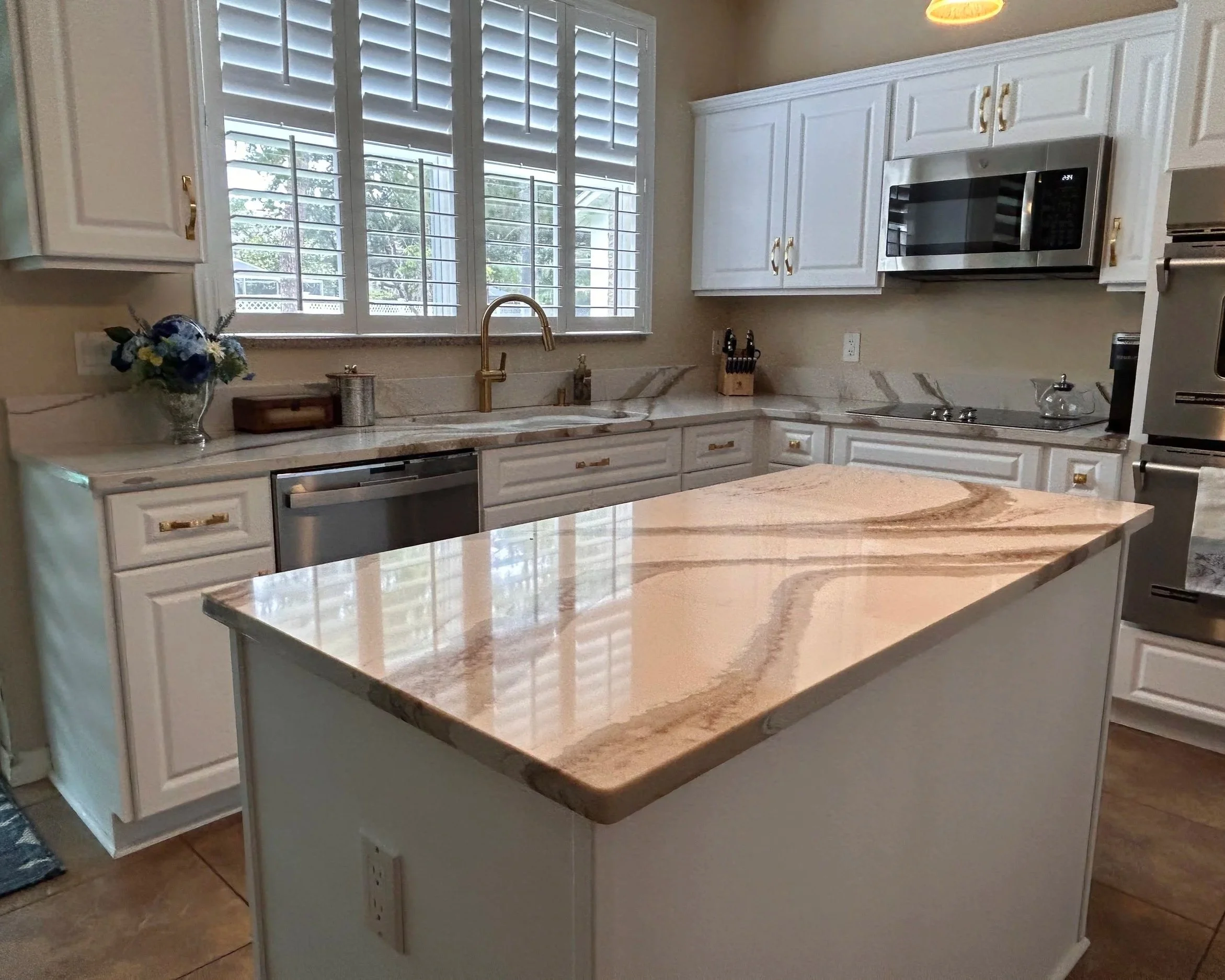 A kitchen with white cabinetry, a marble countertop, a stainless steel microwave, and a window with white plantation shutters. There is a vase of flowers on the counter and various kitchen appliances.