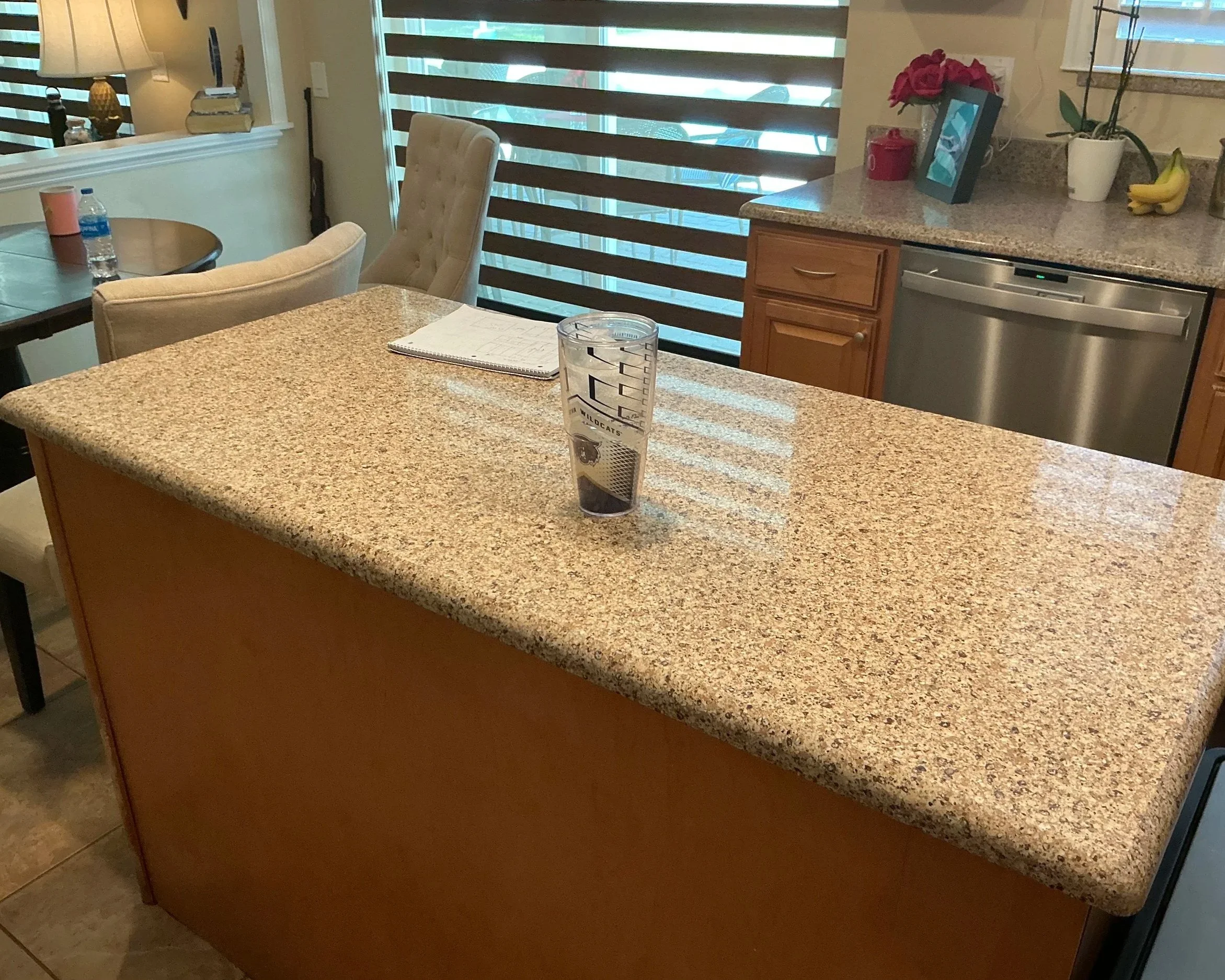 A kitchen with a granite countertop, a beige tufted chair, a counter with a flower pot, bananas, a red container, a picture frame, a water glass with a sports team logo, and a water bottle. A sliding window with striped shades is in the background, and a dining table with chairs is visible to the left.