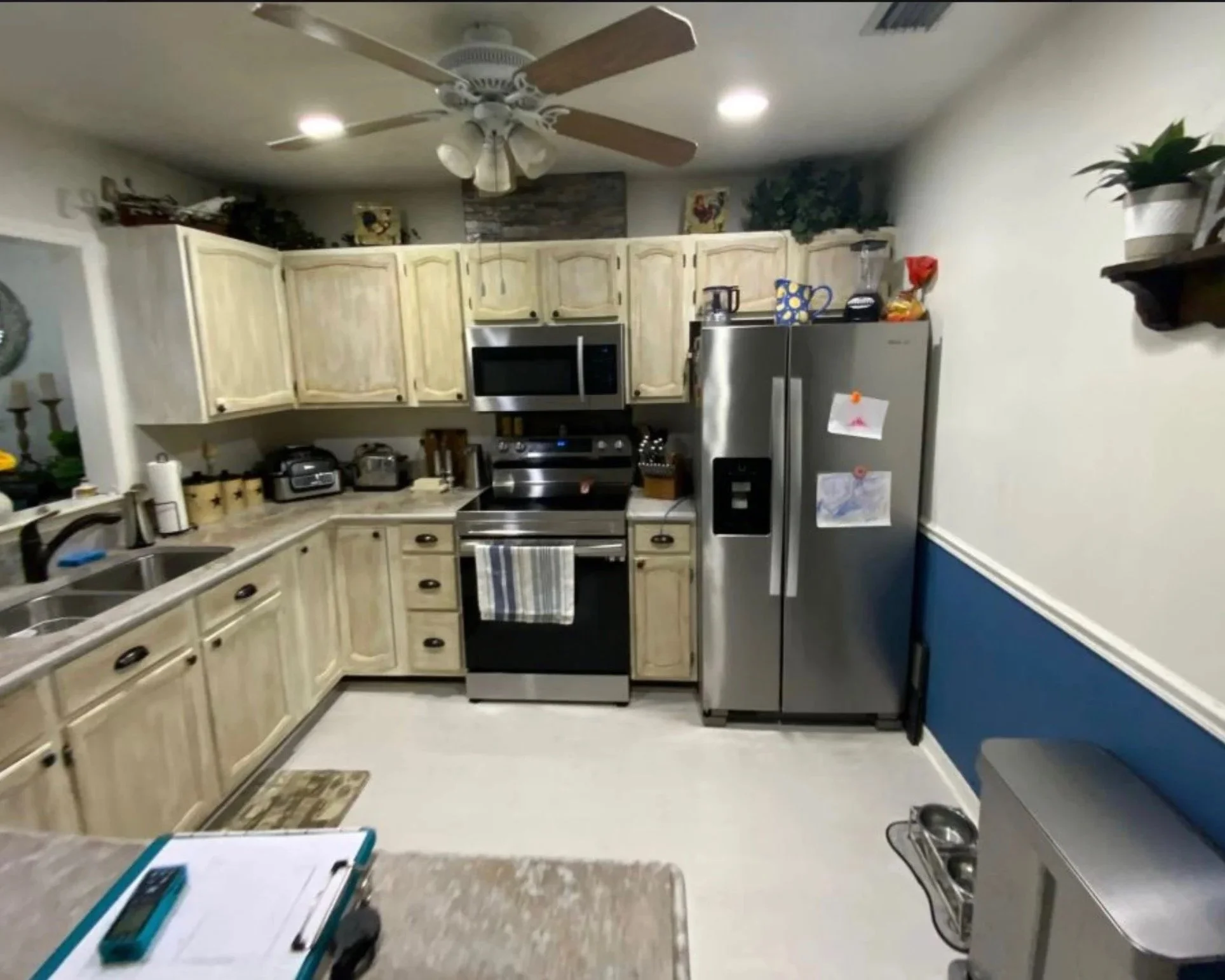 A kitchen with light wood cabinets, stainless steel appliances, including a refrigerator and microwave, and various kitchen items on the counter and shelves. There is a ceiling fan with lights, and a section of countertop with a dish drying rack in the foreground.