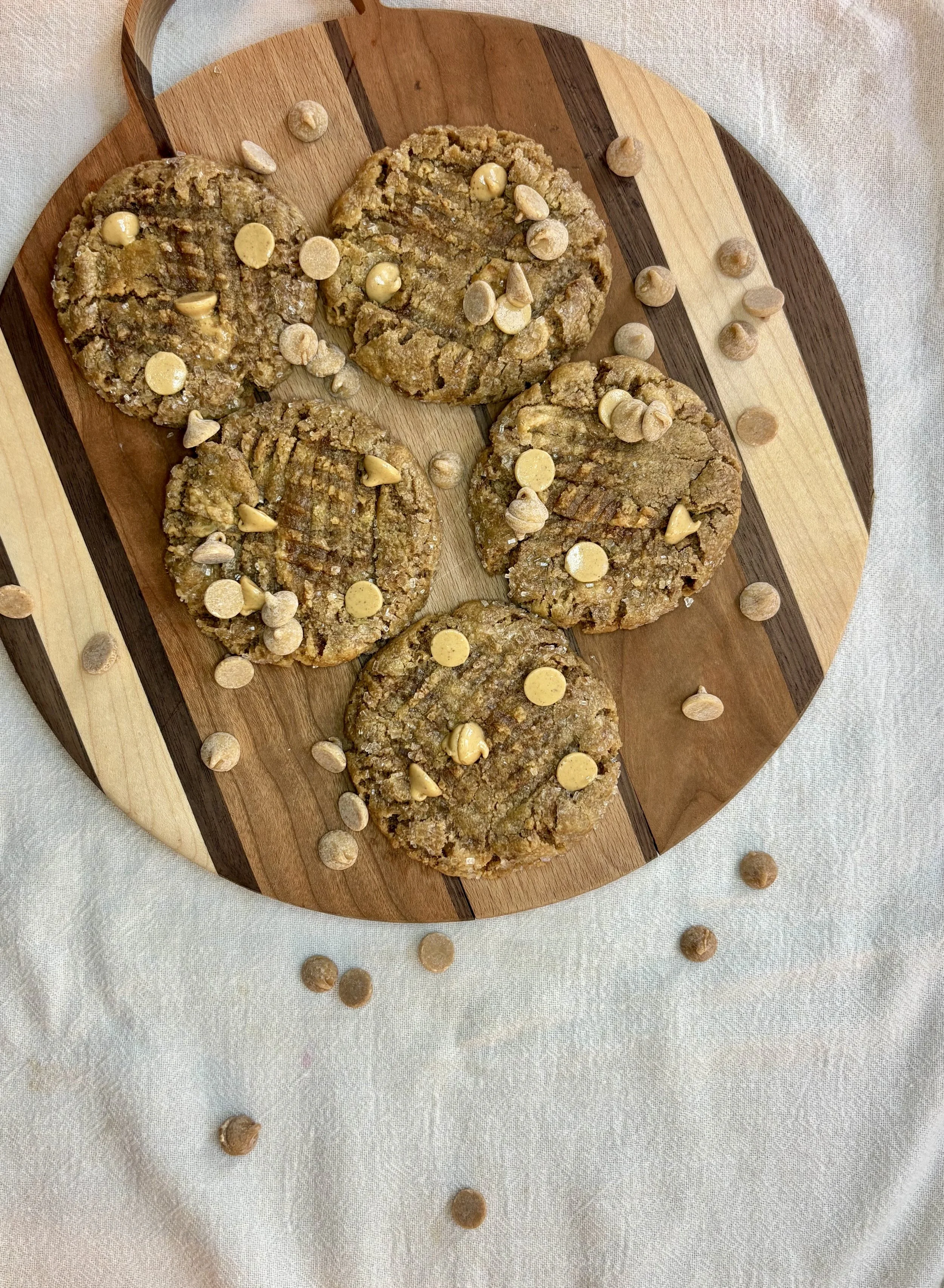 Four oatmeal cookies topped with white chocolate chips and sprinkled with cashew nuts on a round wooden serving board, with extra cashew nuts scattered around on a white tablecloth.