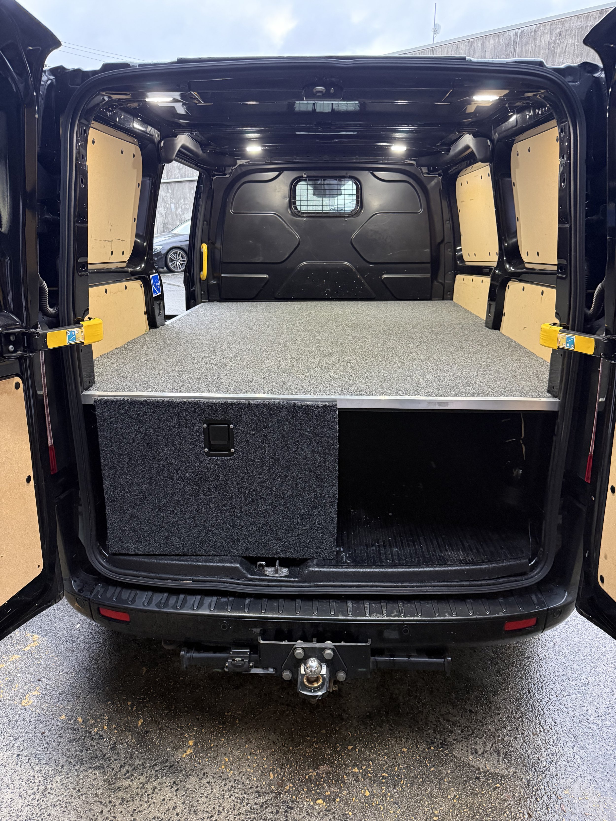 Back of a black utility van with an open rear door revealing a cargo space with a textured gray floor mat and beige side panels, a tow hitch attached to the rear bumper.