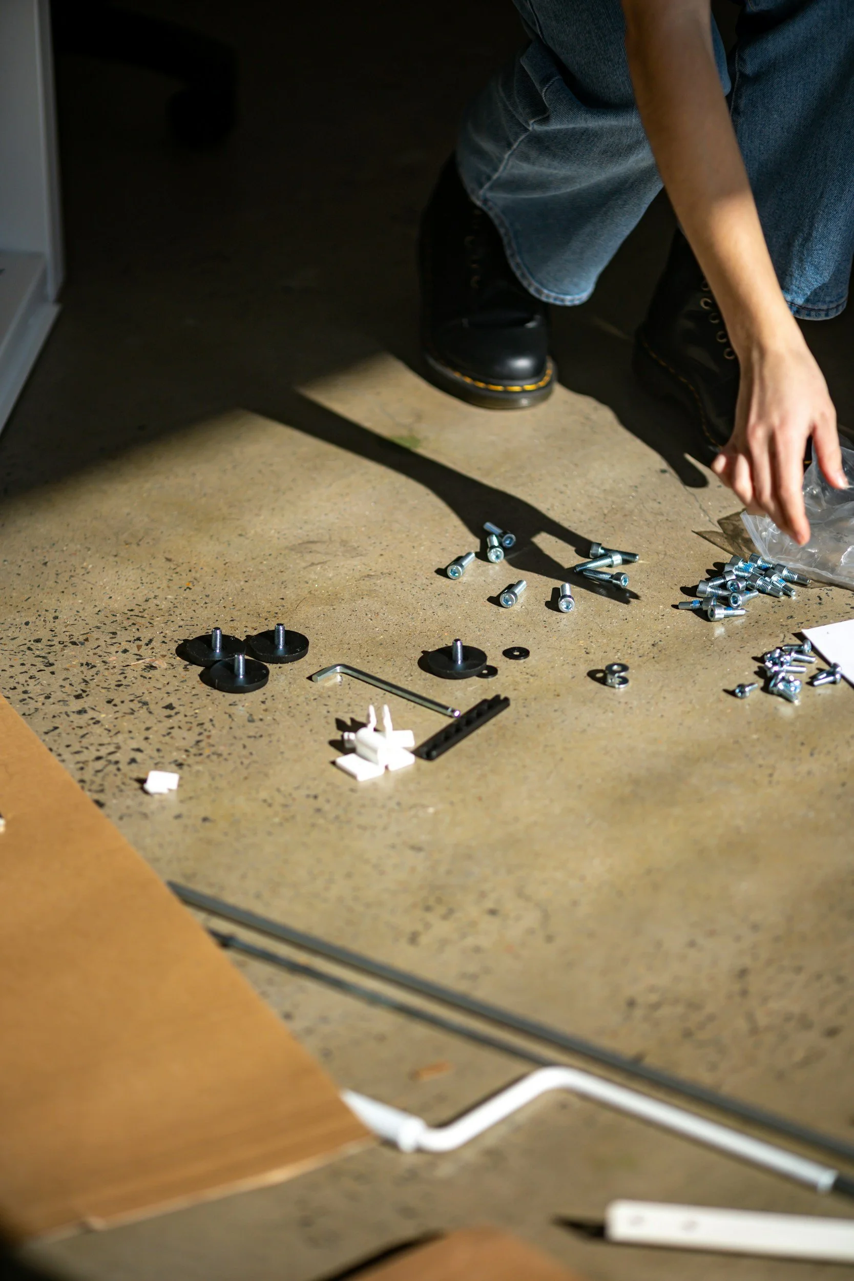 Person assembling or repairing furniture, kneeling on the floor, with various screws and small hardware scattered around.