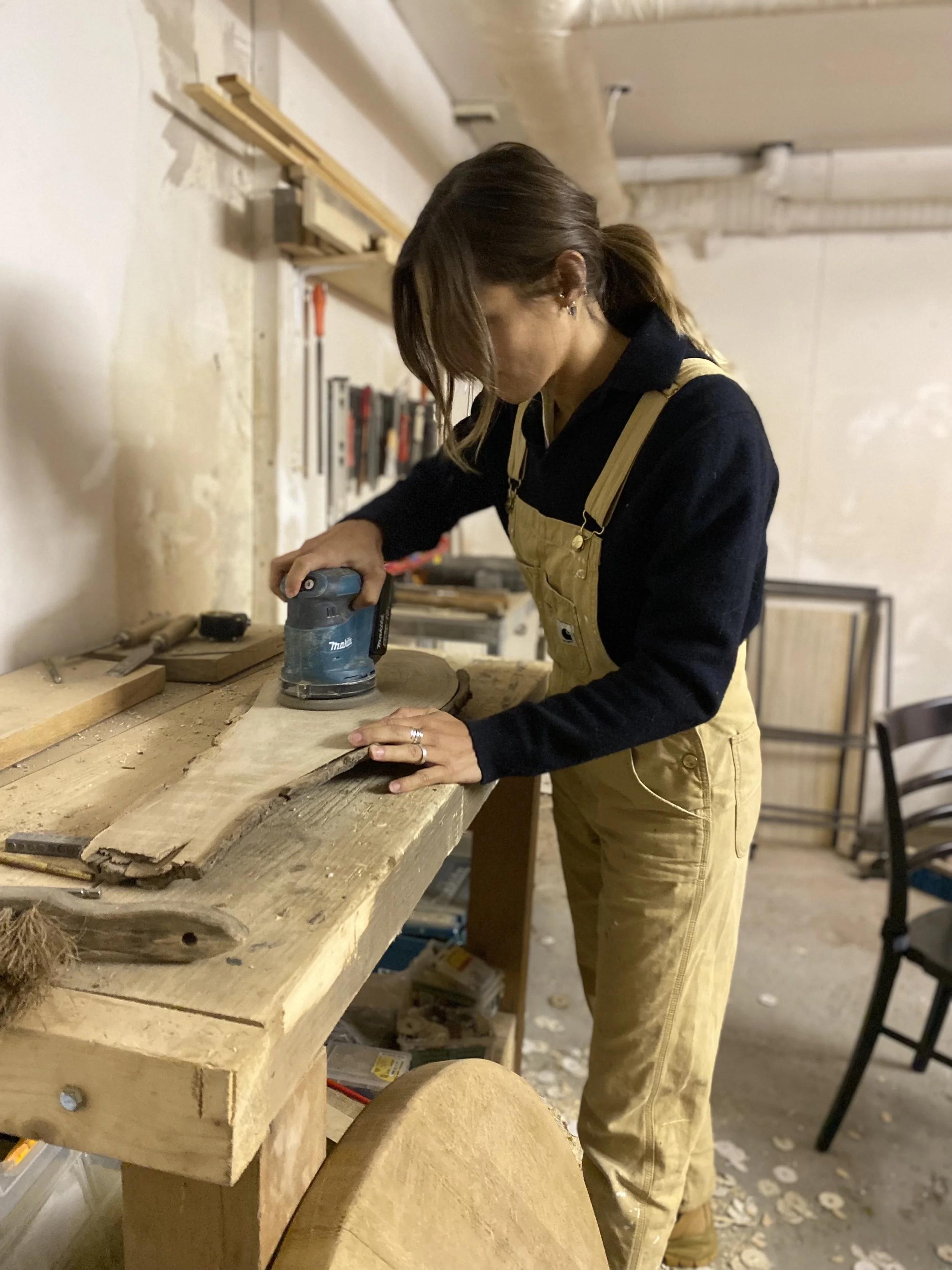 A woman in yellow overalls working on a woodworking project, sanding a large piece of wood with an electric sander in a workshop.