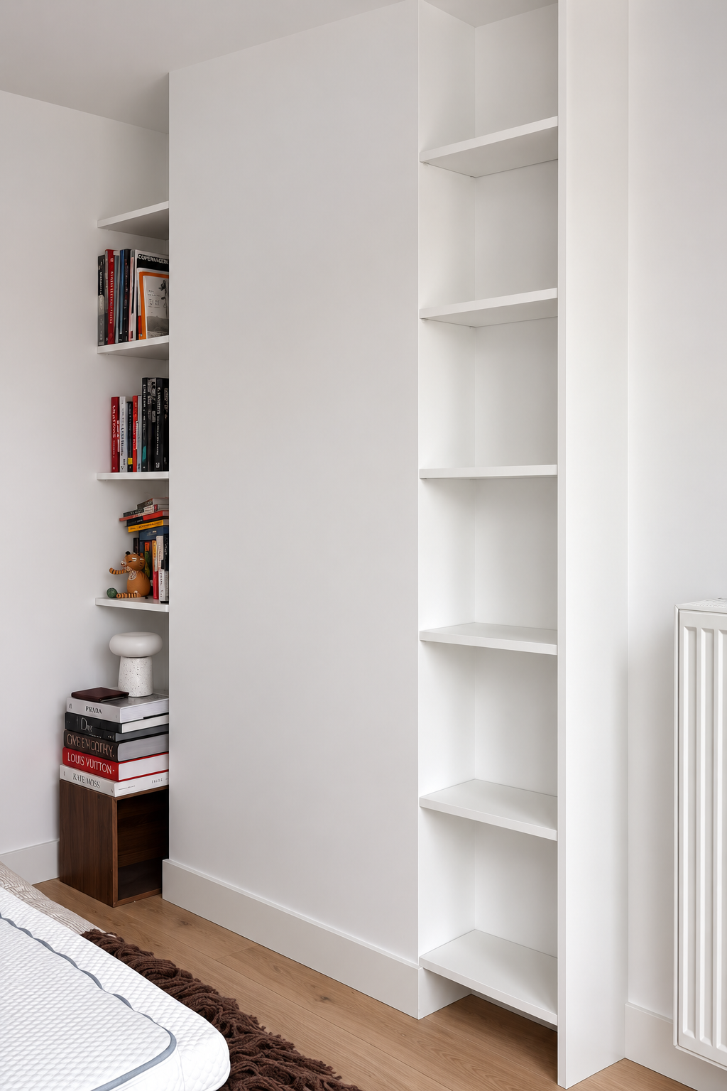 White built-in bookshelves with several books and decorative items, next to a white wall, part of a bedroom with a wooden floor.