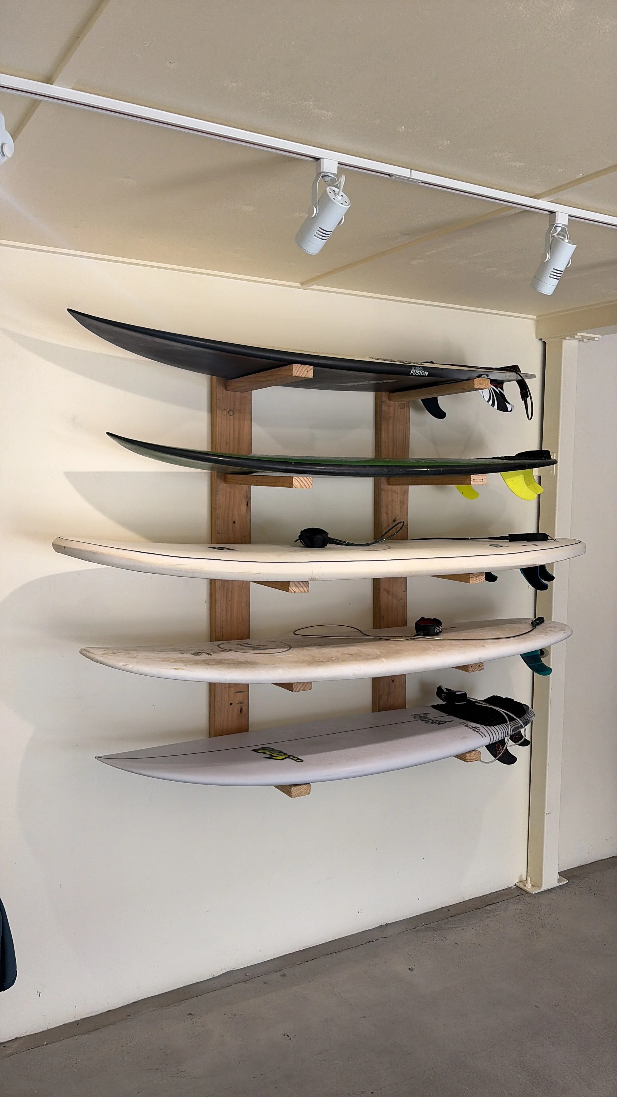 A wall-mounted storage rack holding five surfboards of different sizes and colors, made from recycled timber.