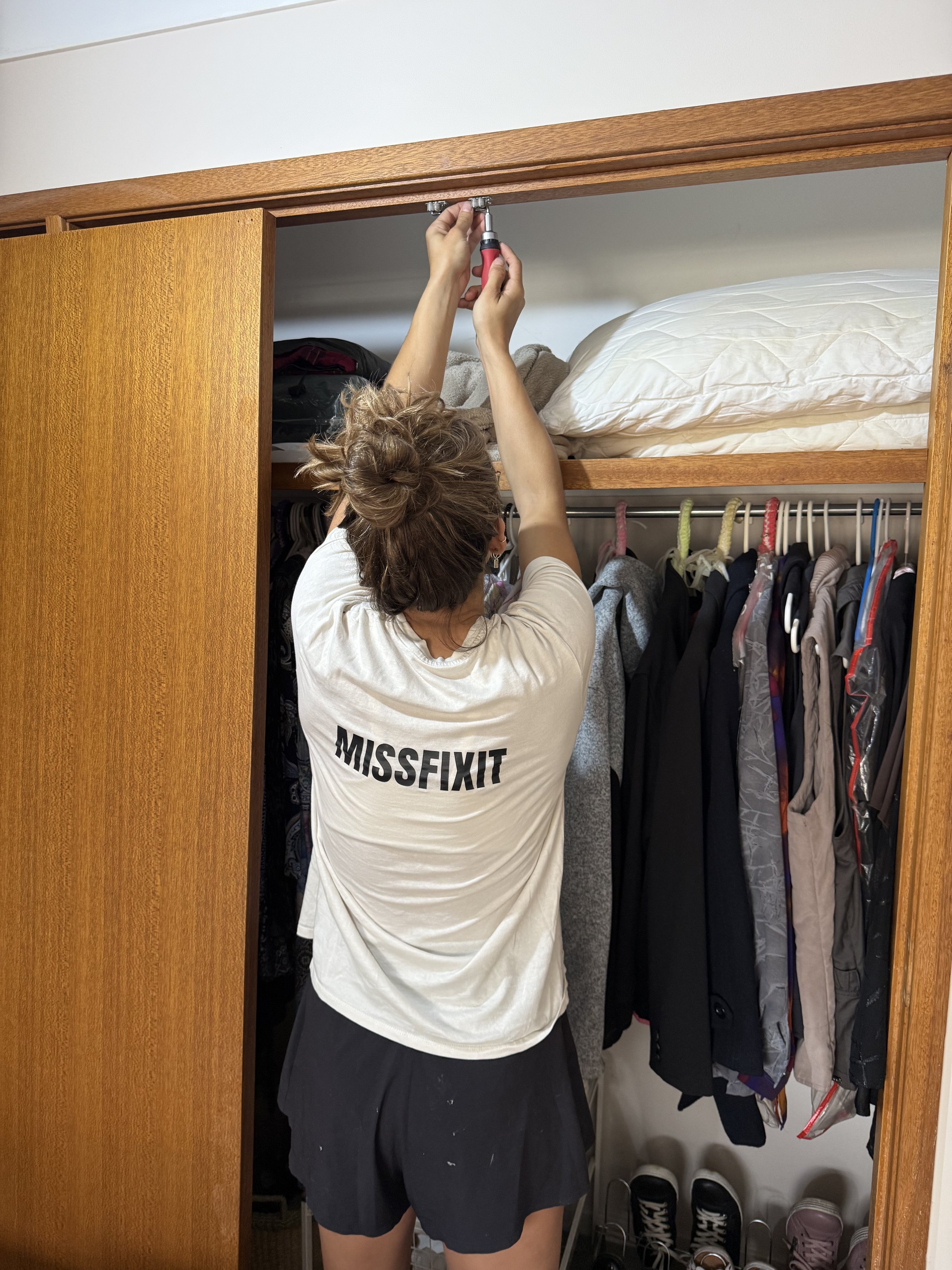 Person with curly hair adjusting a closet shelf using a screwdriver, wearing a white shirt with 'MISSFIXIT' printed on the back, black shorts, and standing in front of a closet filled with clothes and shoes.