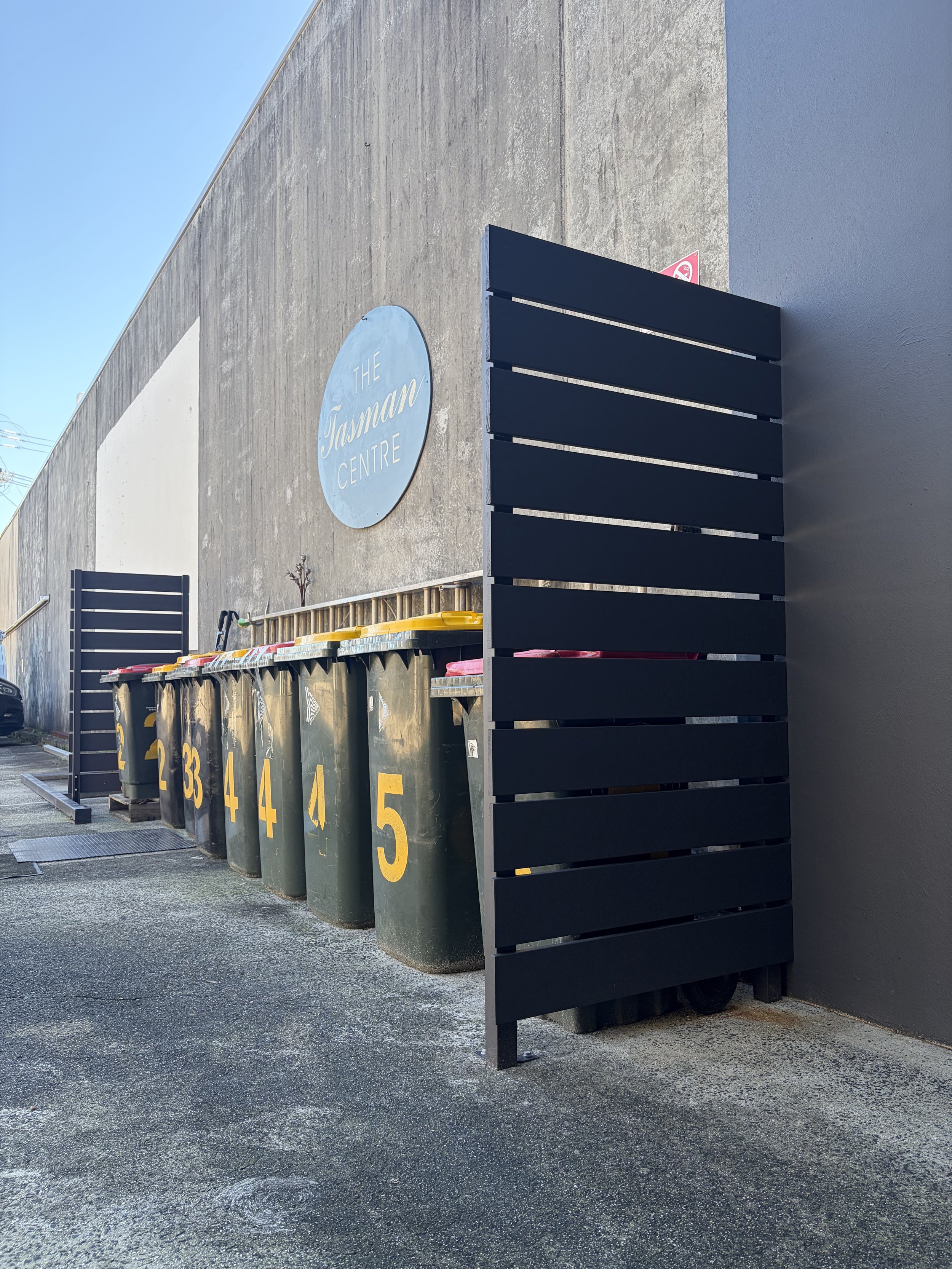 Row of yellow and gray garbage bins with numbers, shielded by two custom built screens standing against a gray wall with a blue sign reading 'The Tasman Centre'.