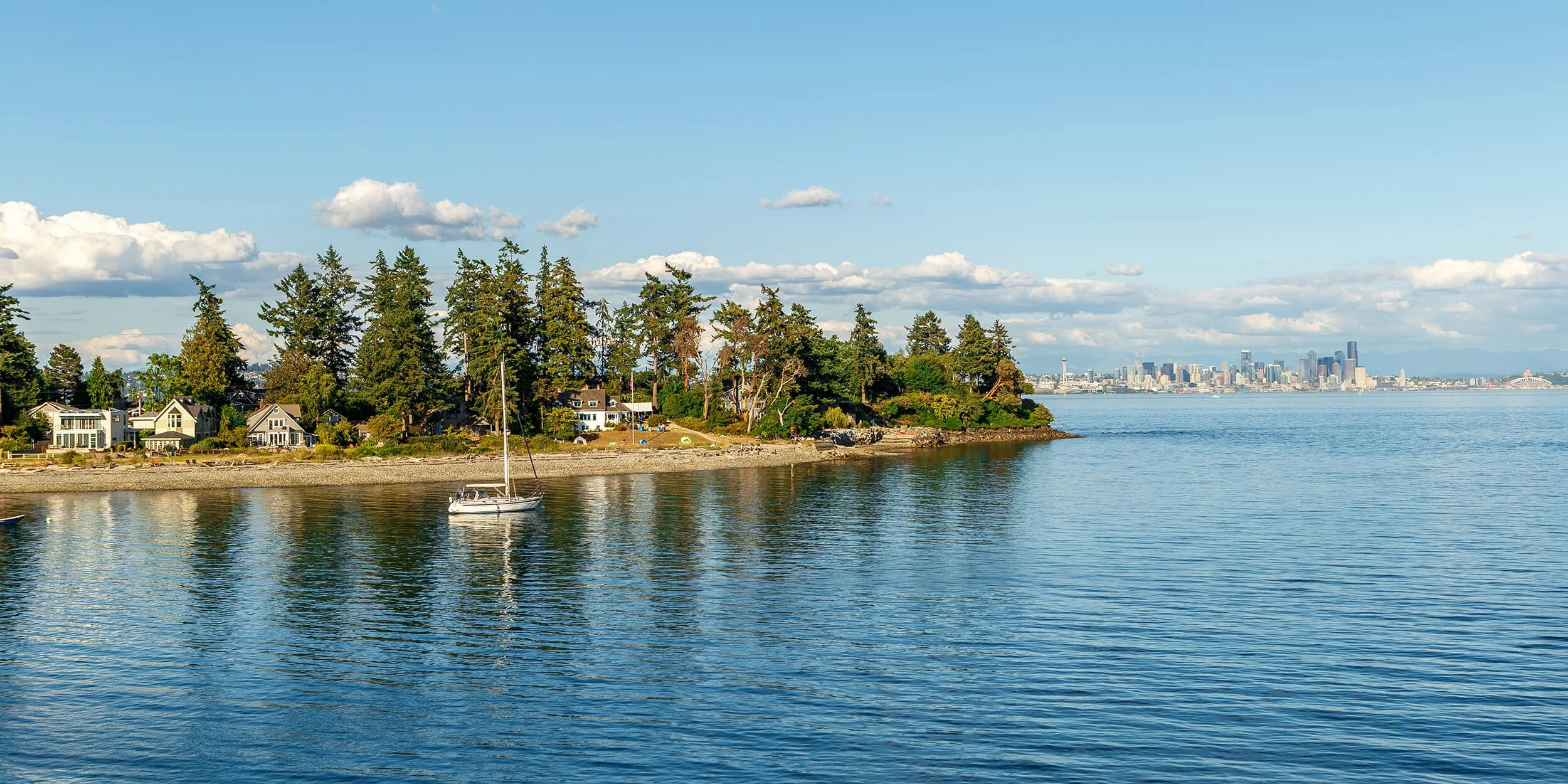 Bainbridge Island waterfront scene featuring houses along the shore, tall trees, a sailboat on the water, and a city skyline in the distance under a partly cloudy sky.