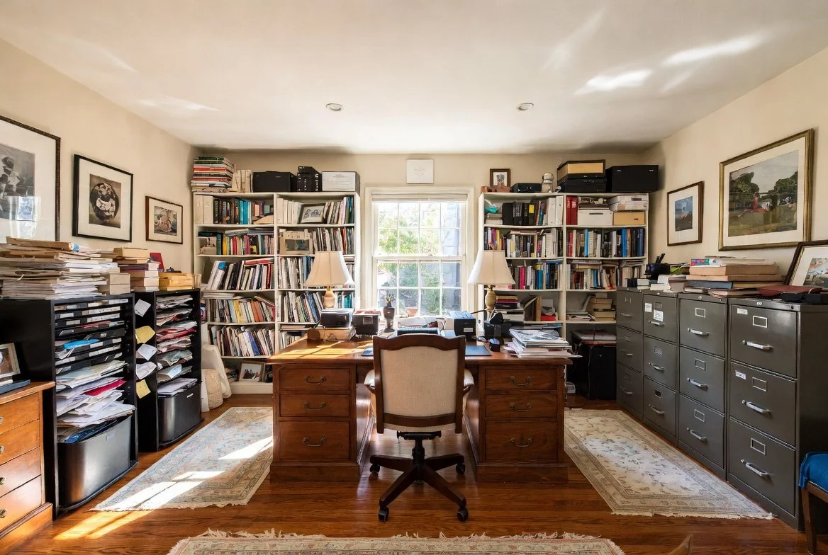 A cluttered home office with a wooden desk, chair, and two bookshelves filled with books and files. There are stacks of papers on black filing cabinets and next to the desk. A window at the back lets in natural light, illuminating the room.