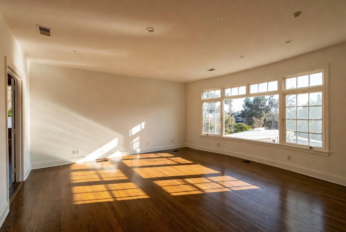 Empty living room with large front window letting in sunlight, hardwood floors, white walls, and a doorway to another room.