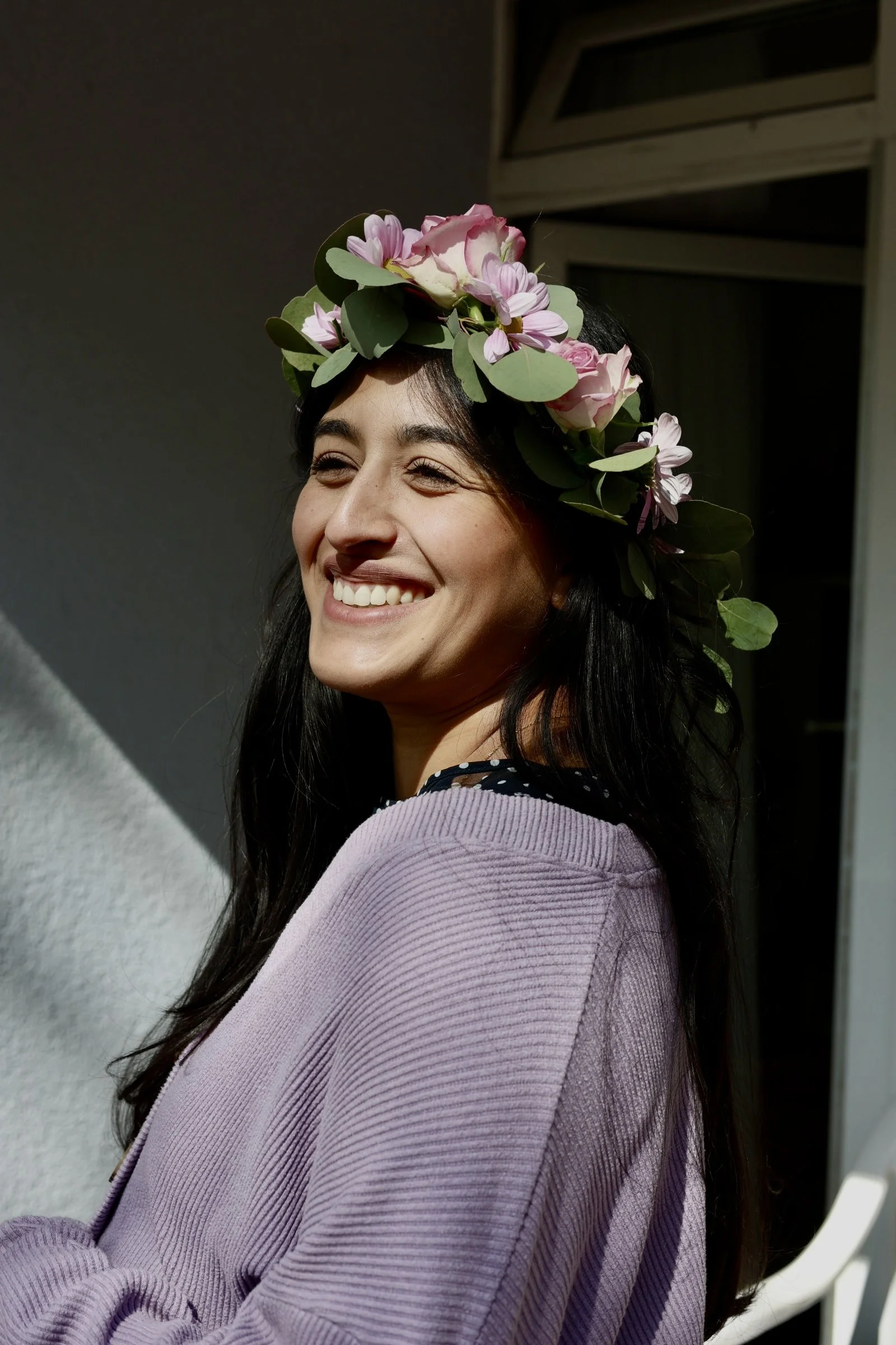 Lächelnde junge Frau mit dunklen Haaren, trägt einen Blumenkranz, besteht aus rosa Rosen und grünen Blättern, trägt einen lavendelfarbenen Pullover, steht vor einem dunklen und hellen Hintergrund, scheint im Sonnenlicht zu stehen.