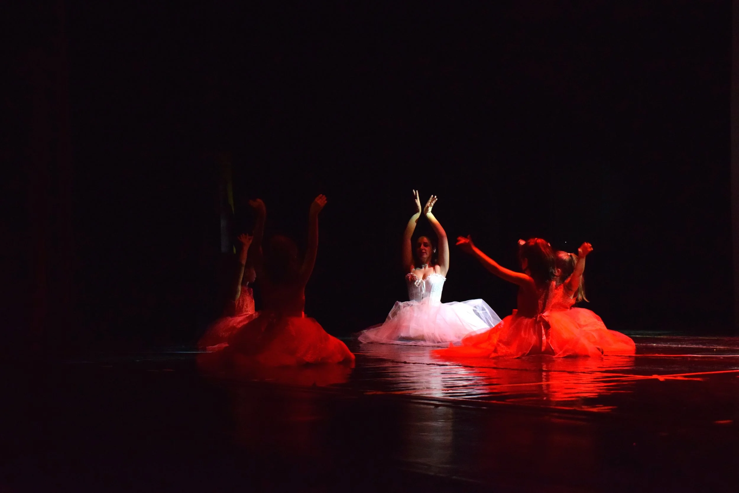 Ballerine in costume bianco e rosa con paillettes, circondate da altre ballerine in abiti arancioni con gonne ampie, tutte che si trovano su un palco con luci rosse e nero.