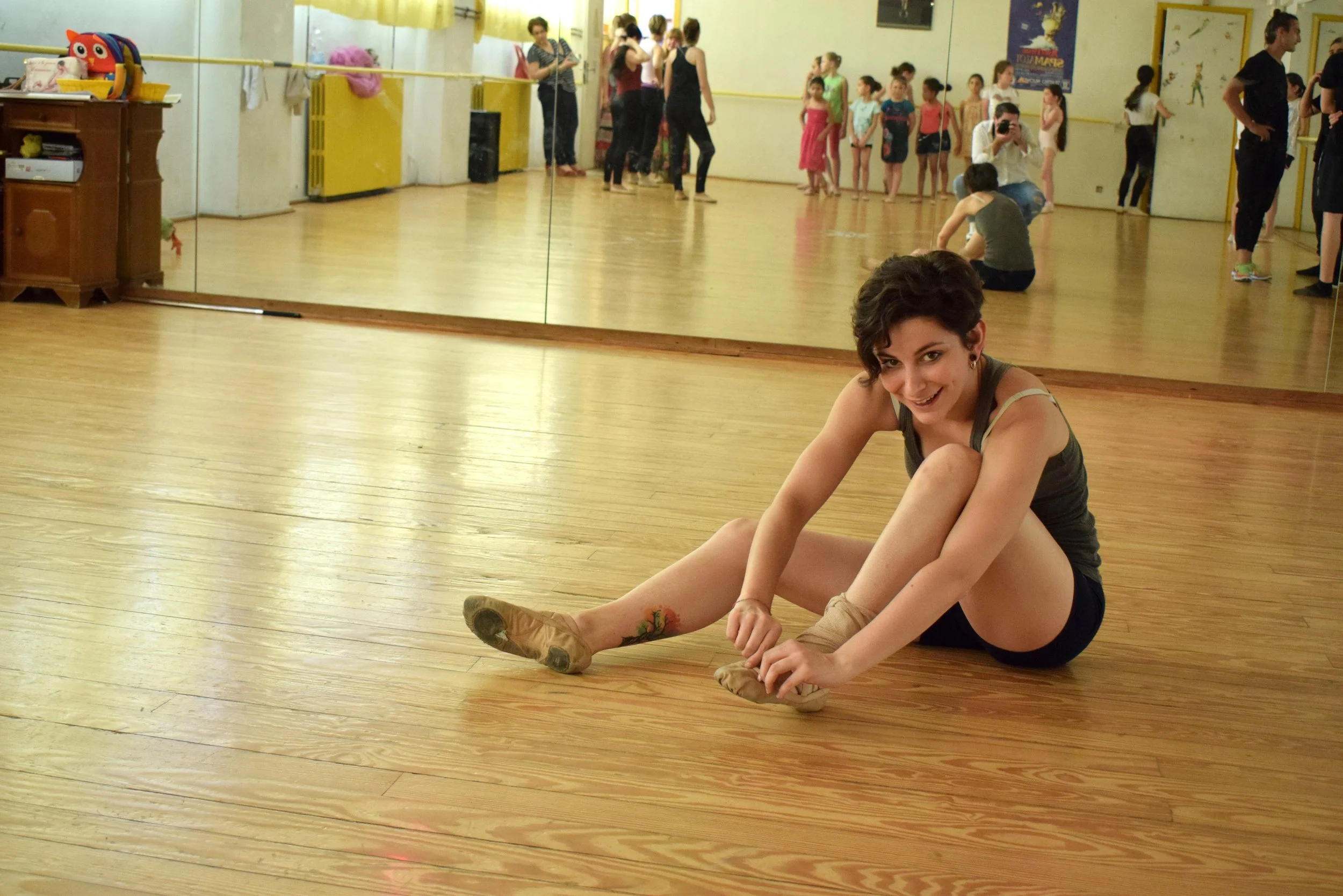 Una donna seduta sul pavimento di legno di uno studio di danza, con una gamba estesa e l'altra piegata, sorridente e intenta ad allacciare le scarpe da balletto. Sullo sfondo, riflesso nel grande specchio, si vedono ragazze e donne che si preparano o