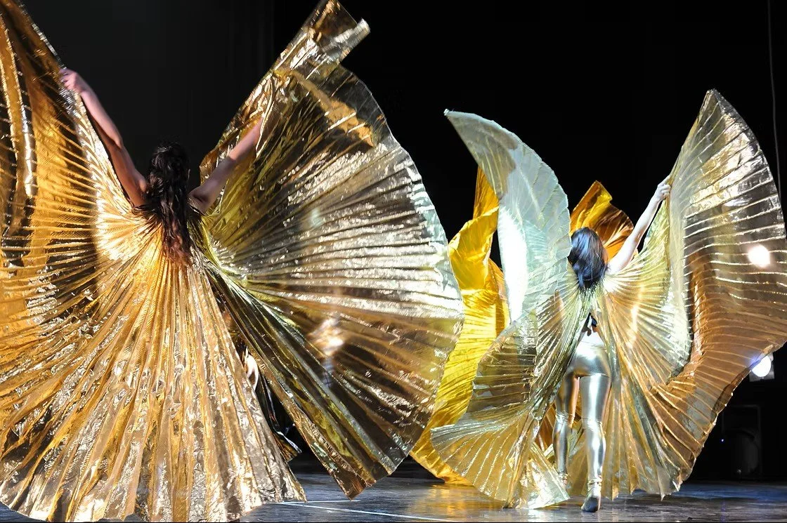 Tre ballerine in costumi dorati con grandi ali di tessuto lucido durante una performance sul palco.