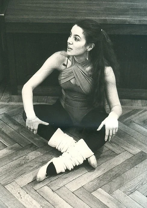 Una donna seduta sul pavimento di legno, vestita casualmente, con le gambe incrociate e indossando calze buffe e scarpe numerose, guarda di lato e sorride leggermente.