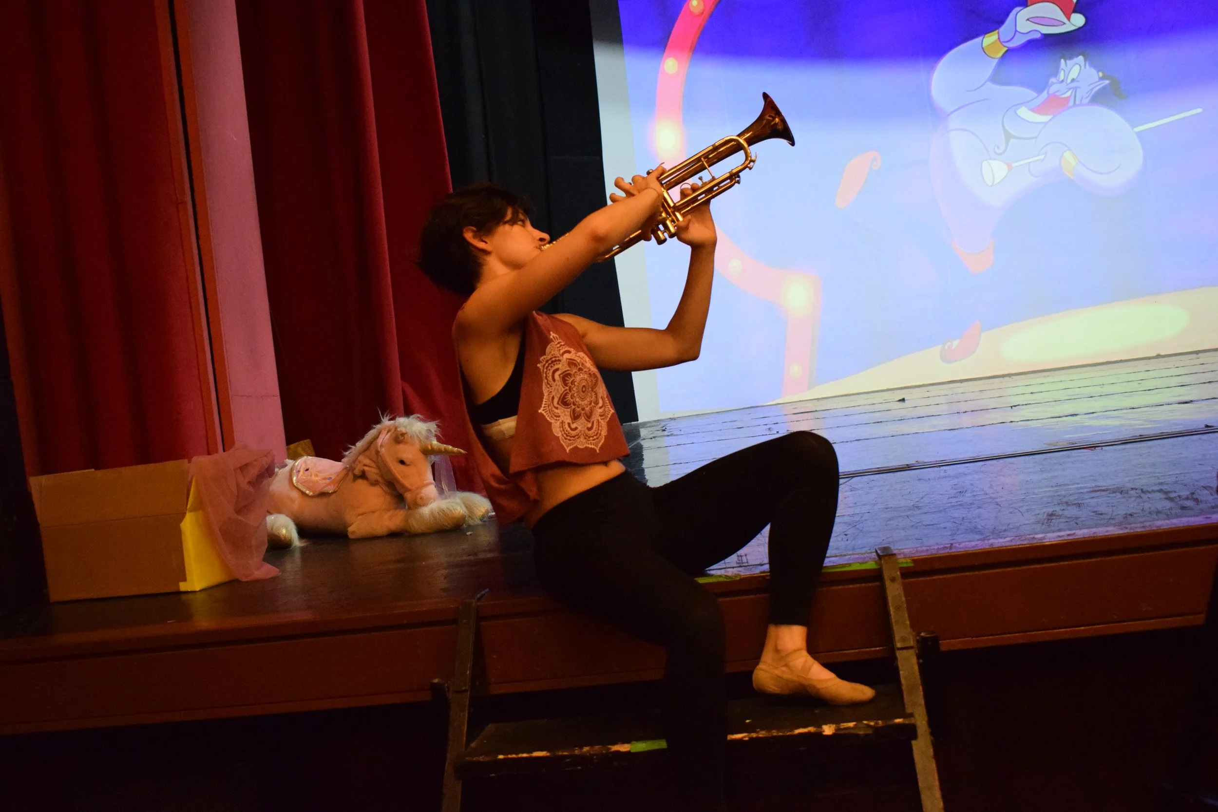 Un bambino suona un sassofono su un palco, con uno sfondo colorato di un cartone animato e un pupazzo di un unicorno sulla scena, seduto accanto a una scatola di cartone.